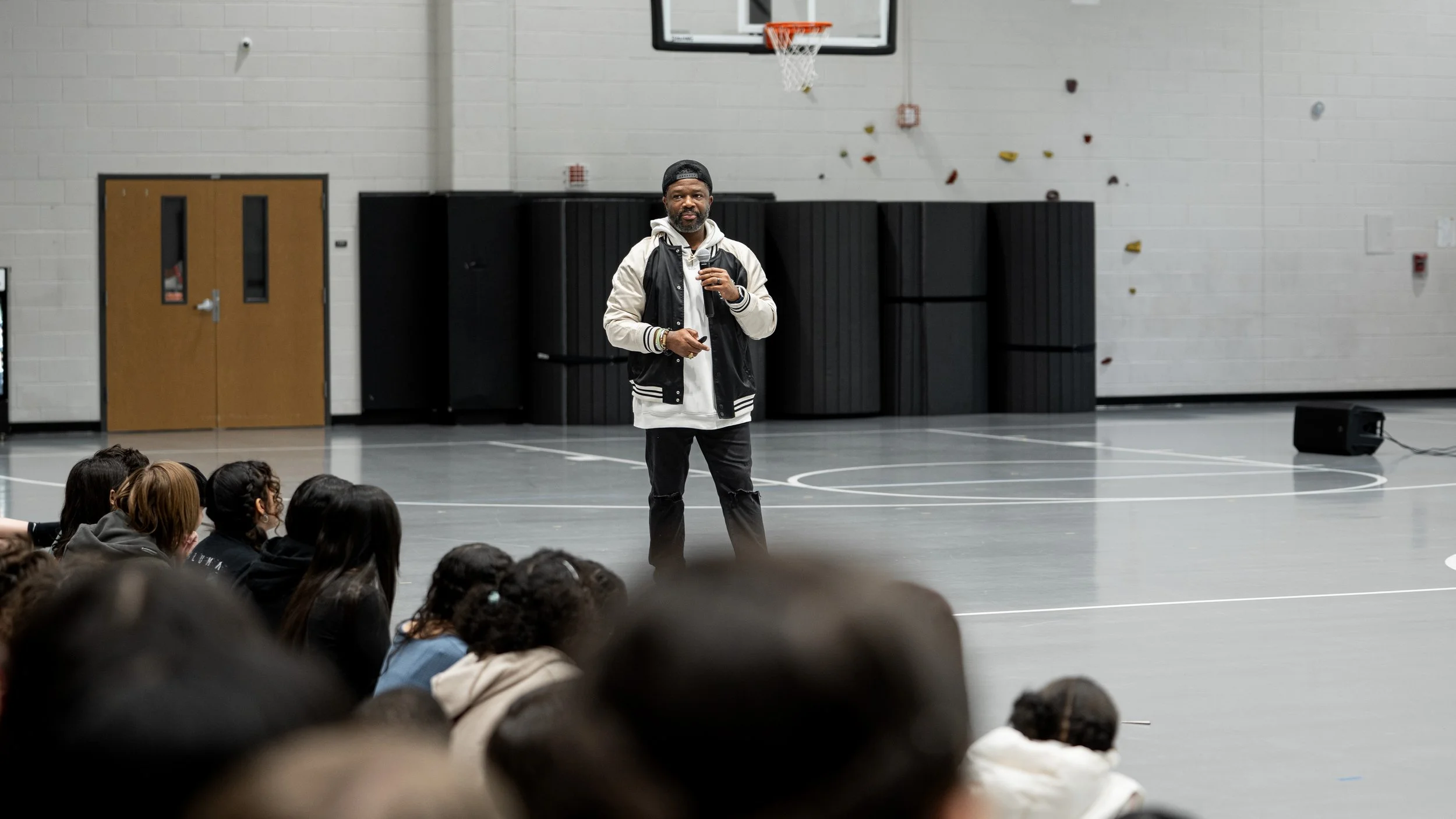 Bobby Gueh with Brothers Making Moves stands in a gymnasium in front of a group of children seated on the floor. He appears to be speaking or giving a presentation while holding a microphone.