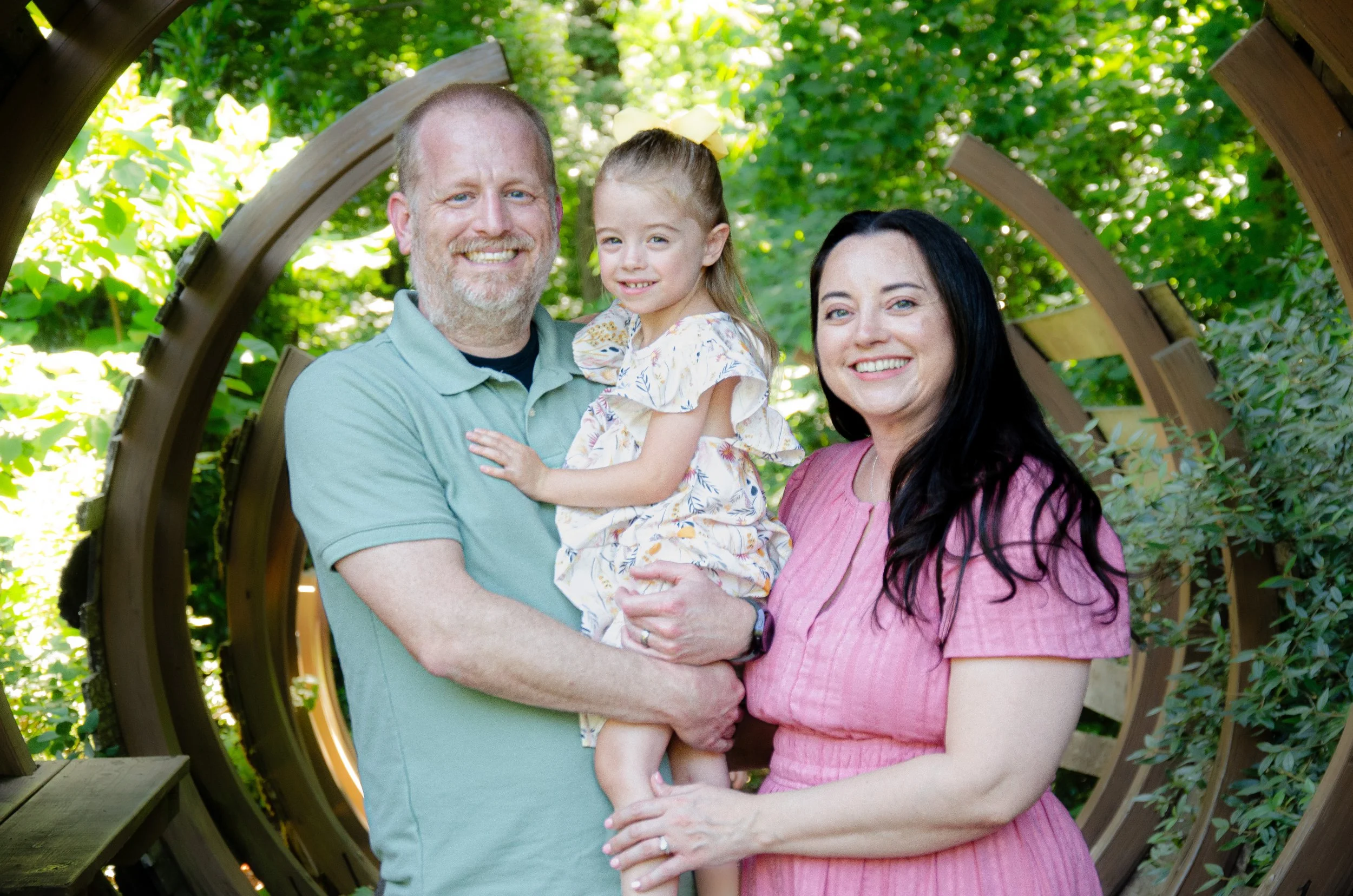 A family of three standing outdoors in a wooden circle structure surrounded by green trees. The man holds a young girl, and a woman stands beside them, all smiling at the camera.