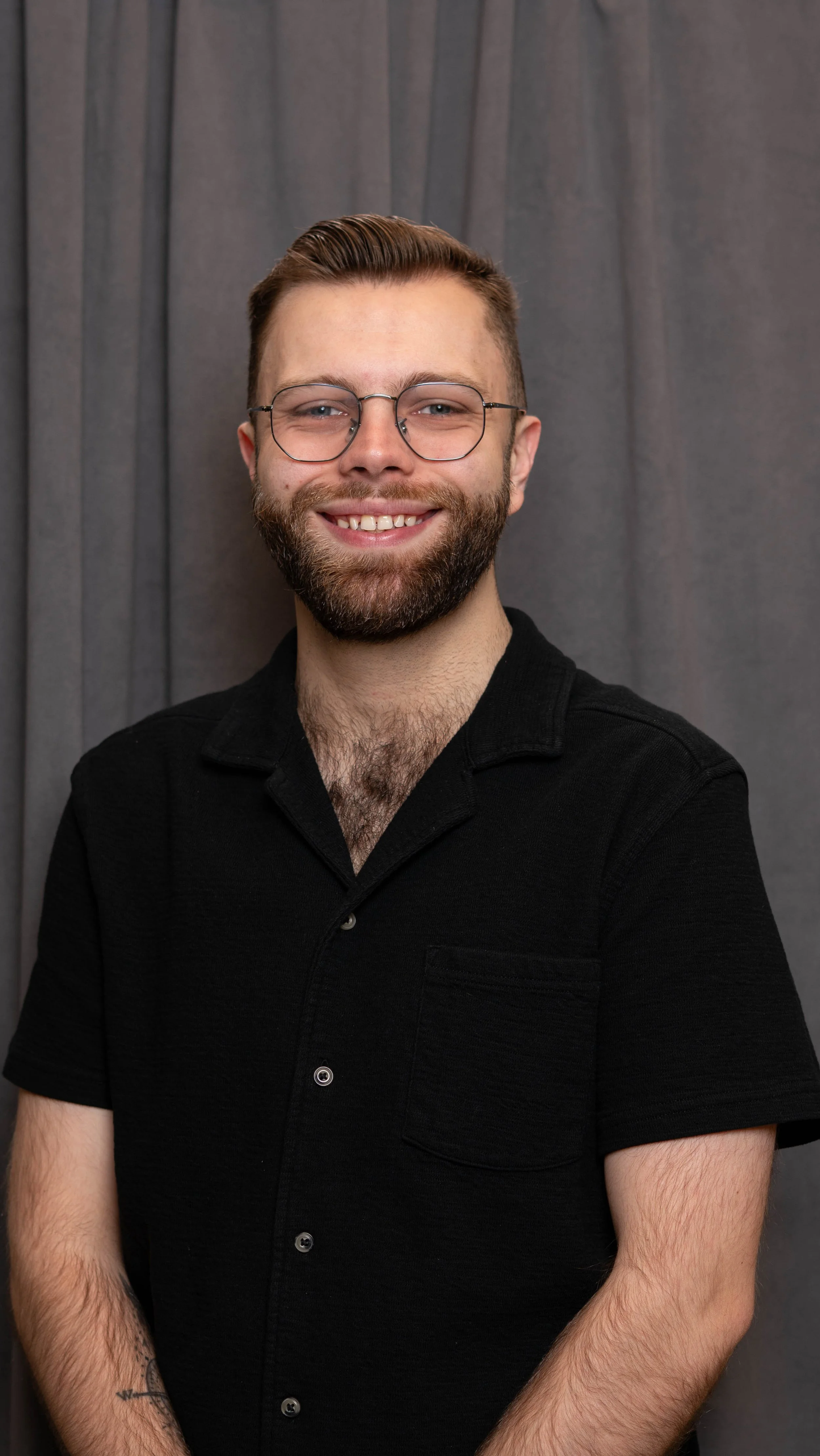 Portrait of a smiling young man with glasses, a beard, and brown hair, wearing a black shirt, standing in front of a grey curtain.