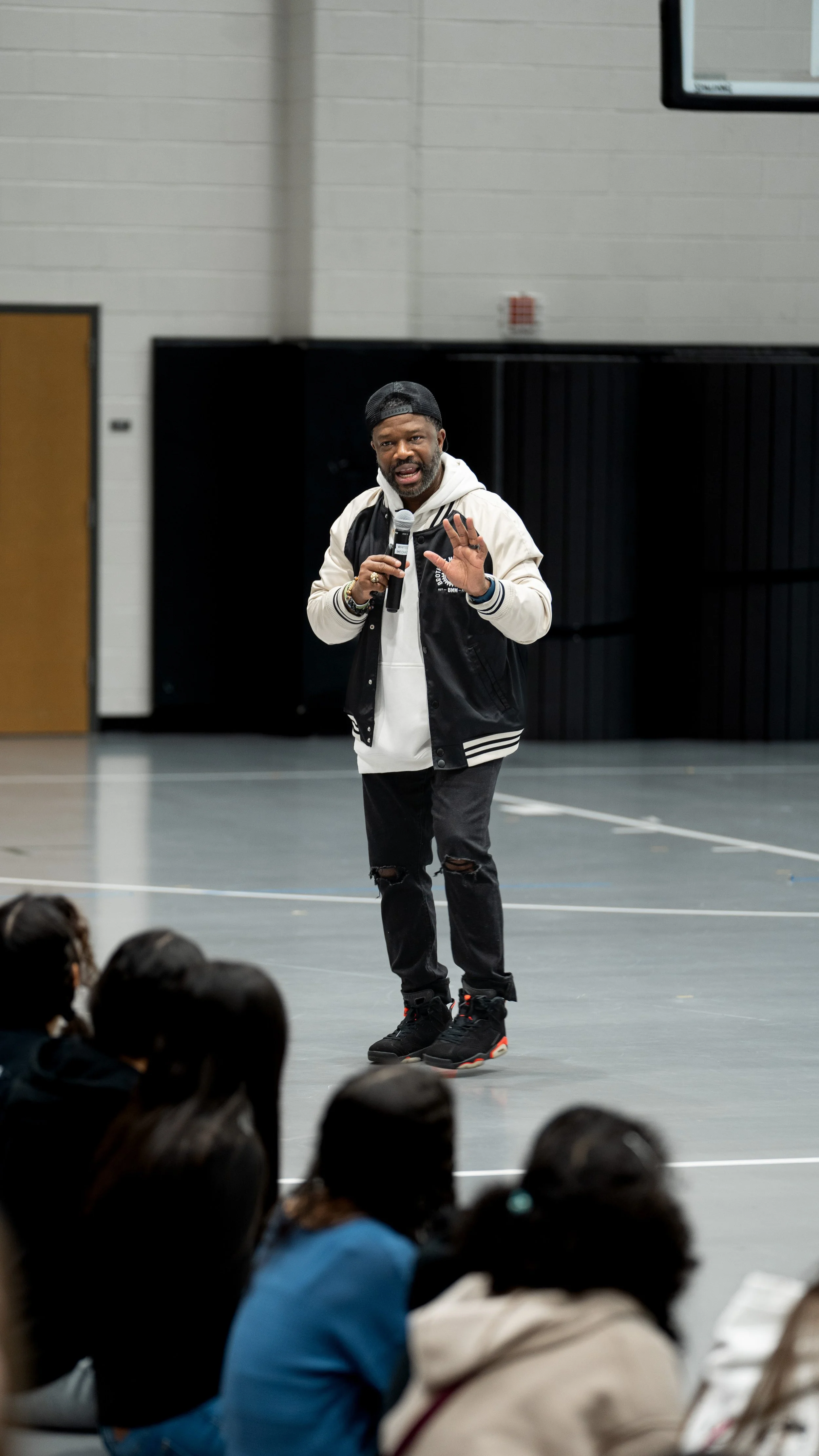 Bobby Gueh with Brothers Making Moves speaking into a microphone in front of a seated audience in a gymnasium.