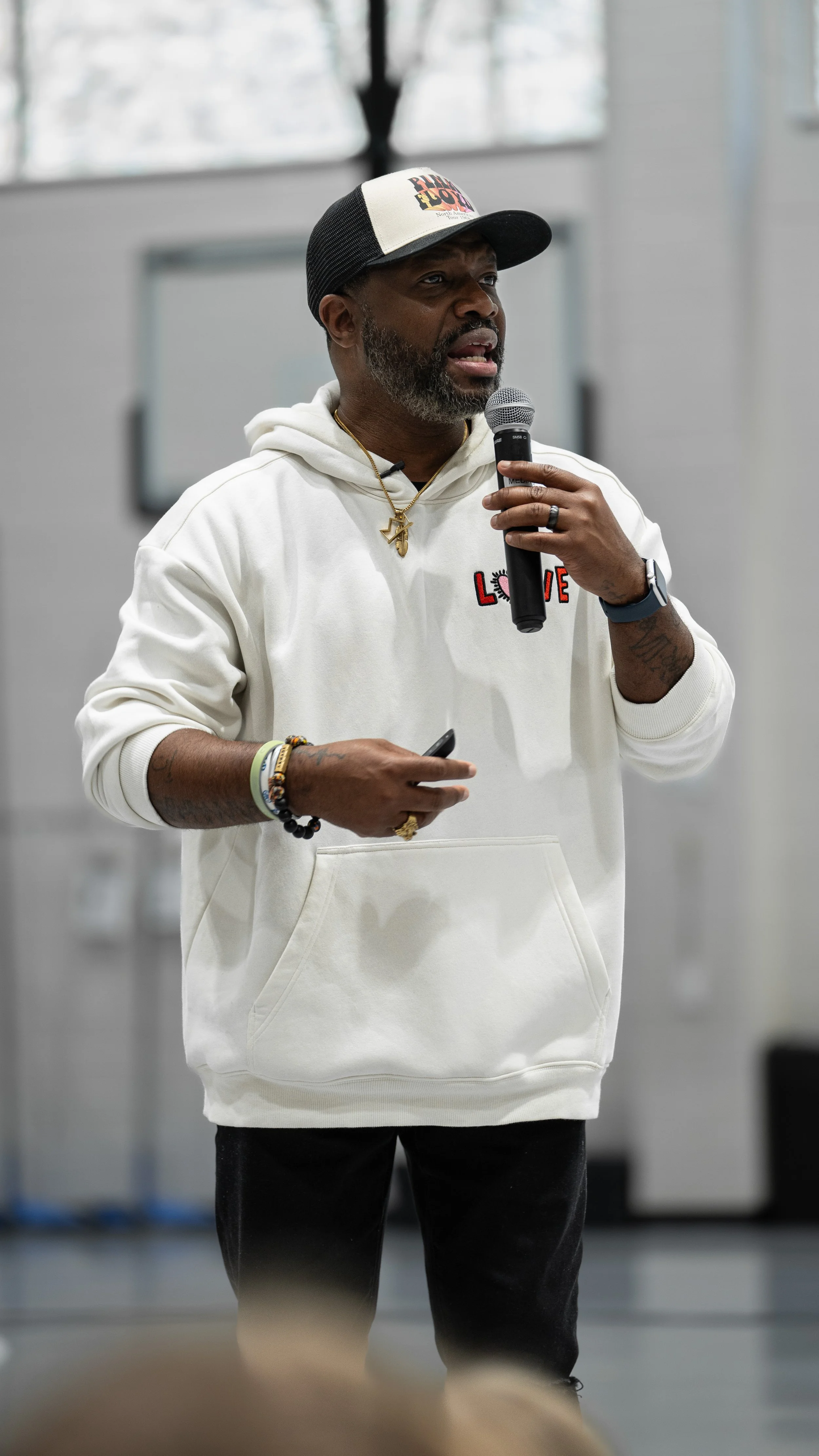 Bobby Gueh with Brothers Making Moves wearing a white hoodie with the word 'LOVE' and a heart on it, holding a microphone, speaking indoors with blurred background.