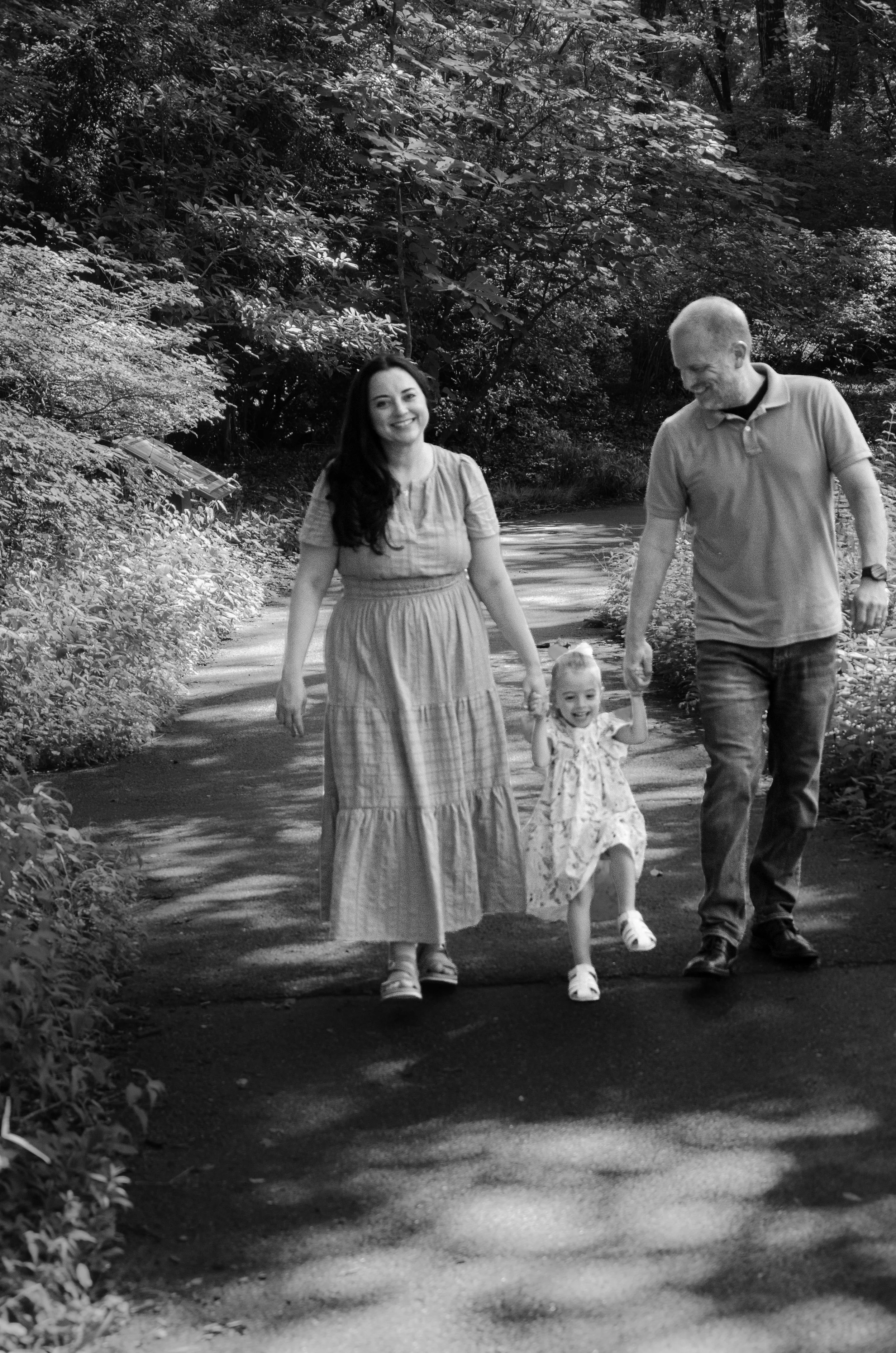 A family of four walking on a shaded forest trail in black and white. The mother is smiling and wearing a long dress, the father is looking down and smiling, the young girl is holding their hands and smiling, and the young boy is walking at the front