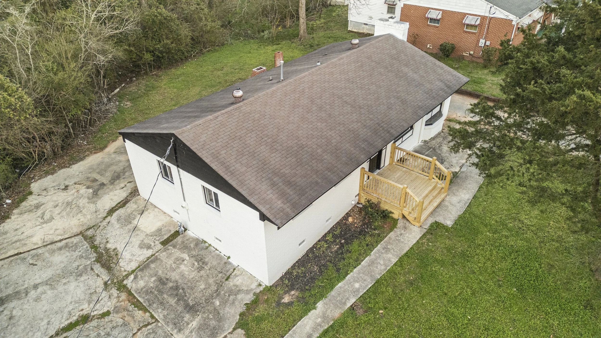 Aerial view of a white house in Athens, GA with a brown shingle roof, a small wooden deck, and surrounding green lawn and trees, with neighboring houses nearby.