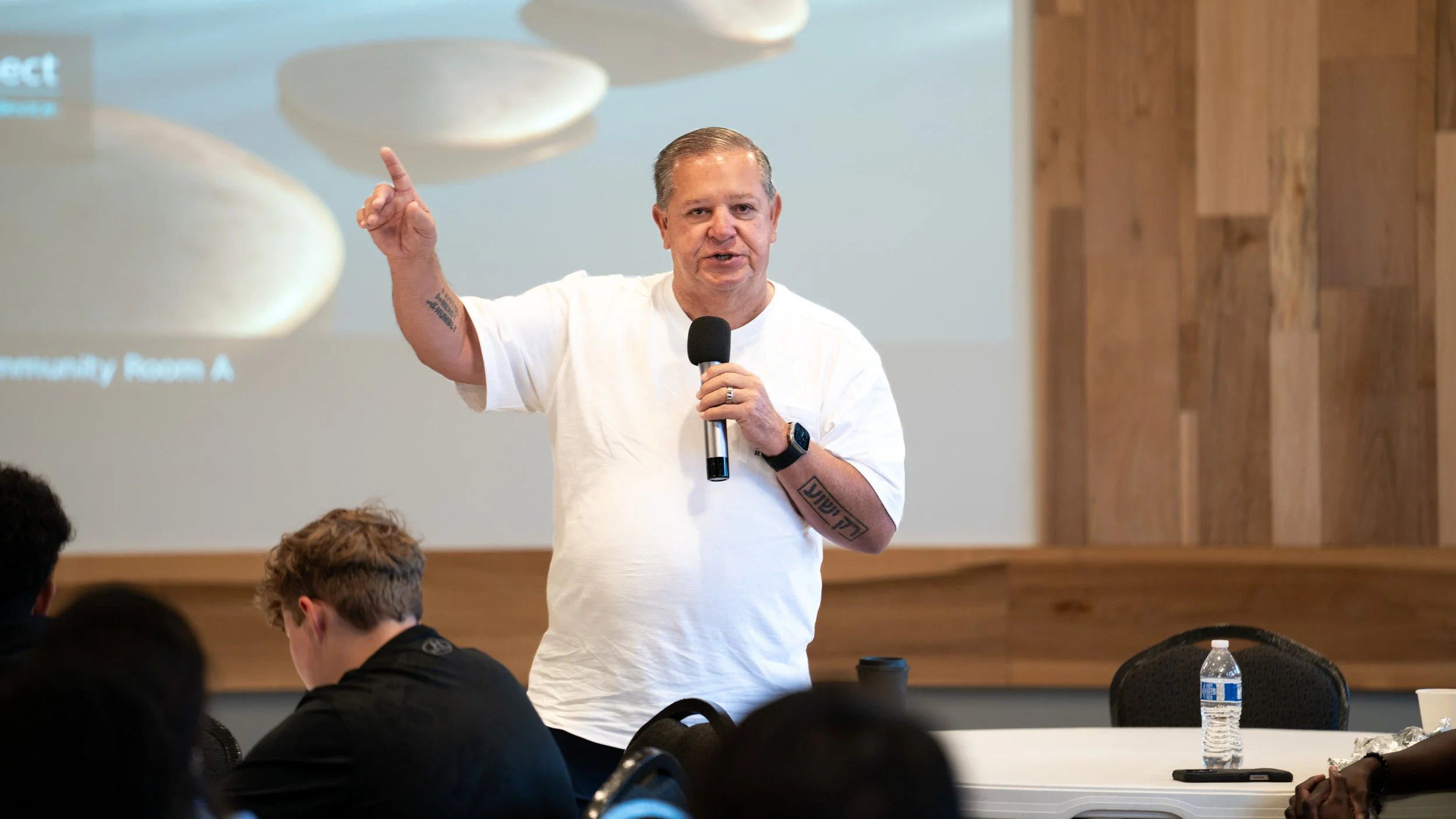 A man with tattoos on his arms speaking into a microphone during a presentation in a room with wooden walls, with people seated at tables listening at Brothers Making Moves.