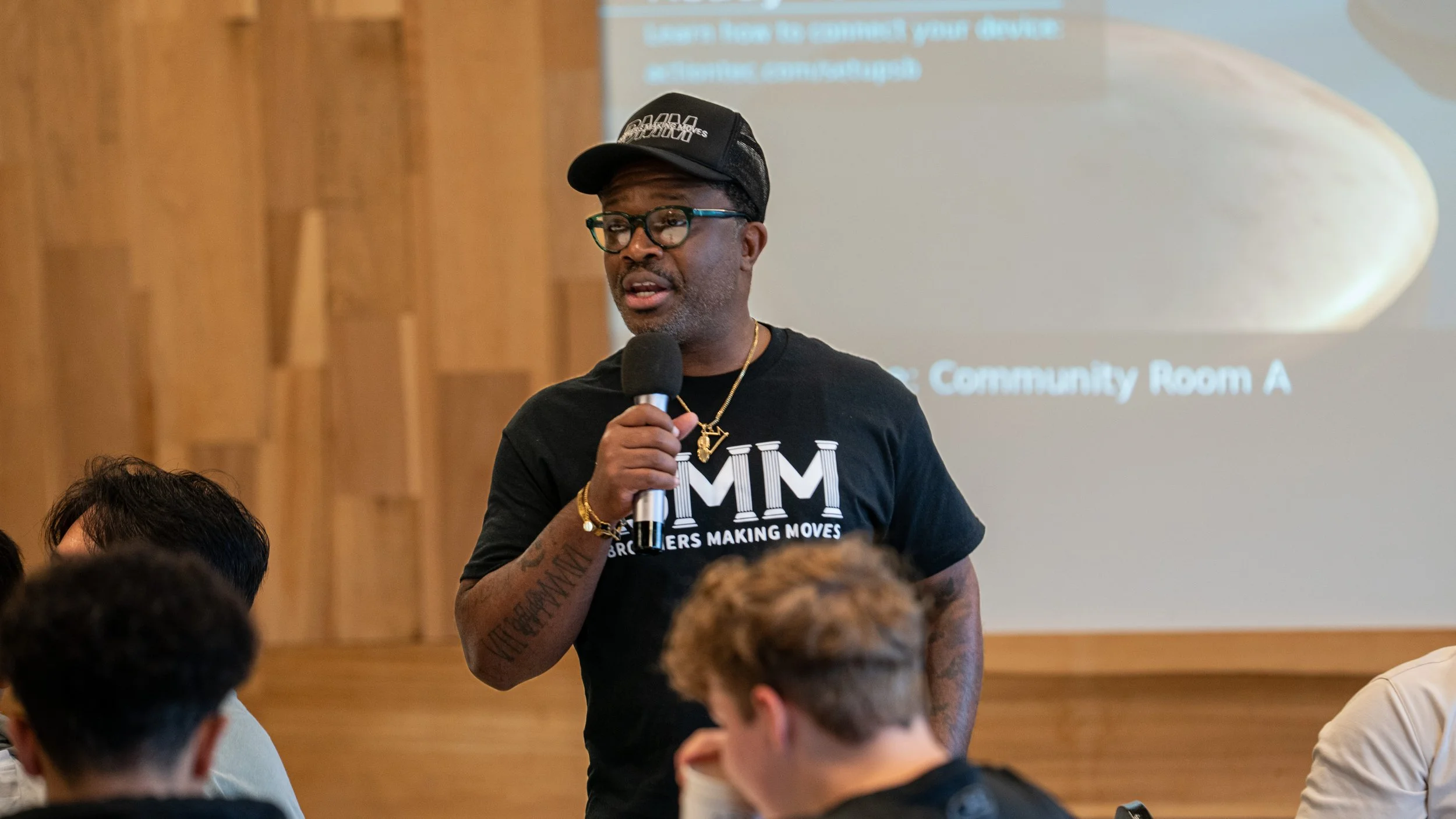 A man with glasses, wearing a black cap and T-shirt with the text 'MIM', speaking into a microphone during a presentation in a room with wooden walls and a projected slide in the background at Brothers Making Moves.