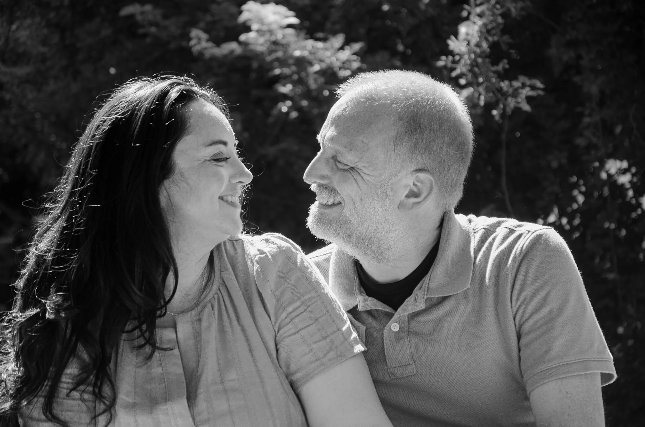A smiling woman with long dark hair looking at a smiling man with short light hair outdoors, with trees in the background.