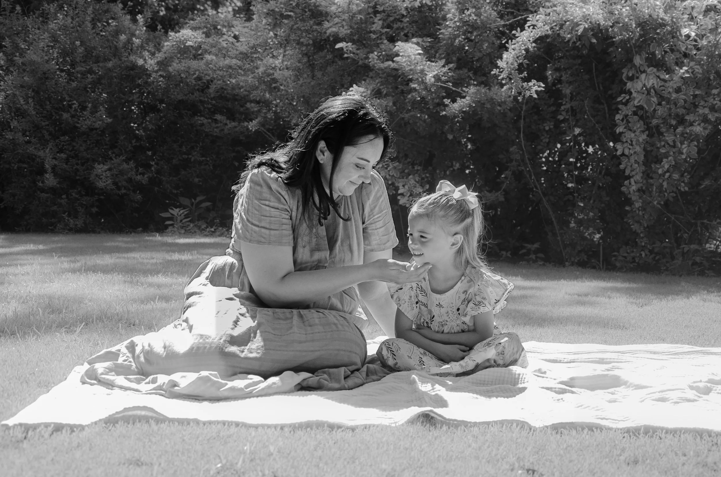A woman and a young girl sitting on a blanket on the grass in a park. The woman is looking at the girl, touching her chin in a gentle manner, and both are smiling. There are trees in the background.