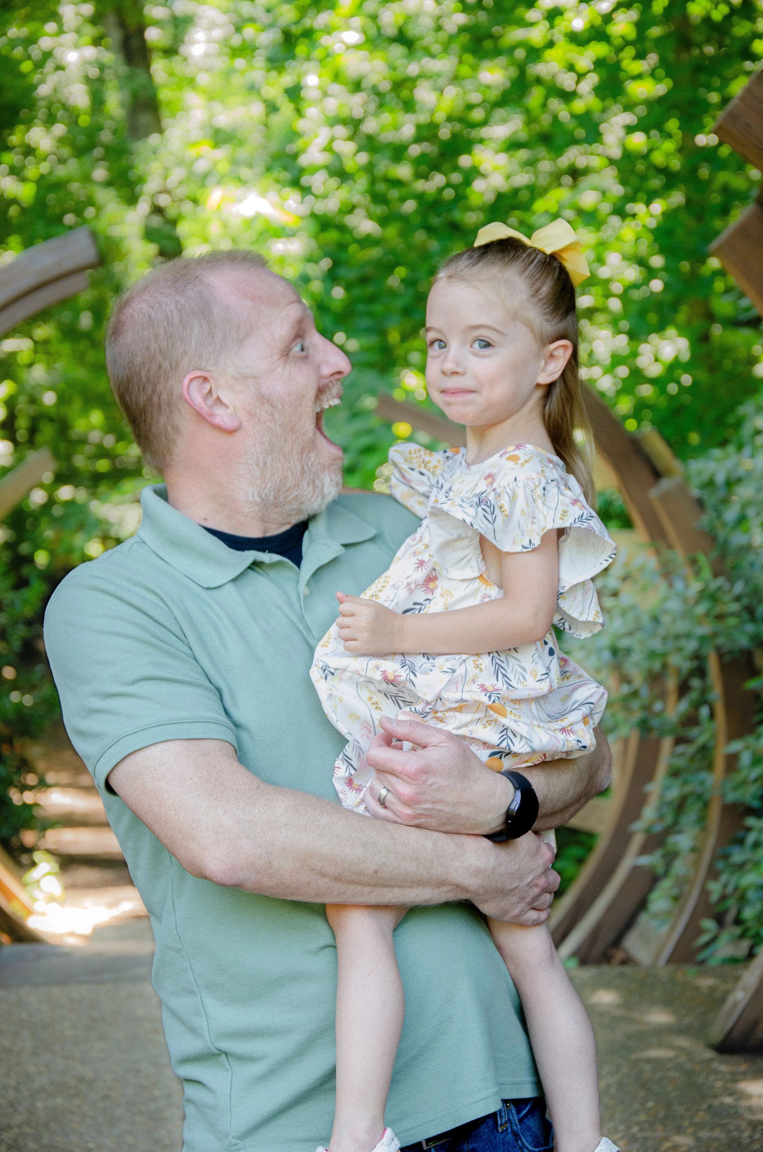A man holding a young girl in a park or wooded area with green trees in the background. The man has short hair and a beard, wearing a light green polo shirt, while the girl has light brown hair tied with a yellow bow and is wearing a floral dress. Th