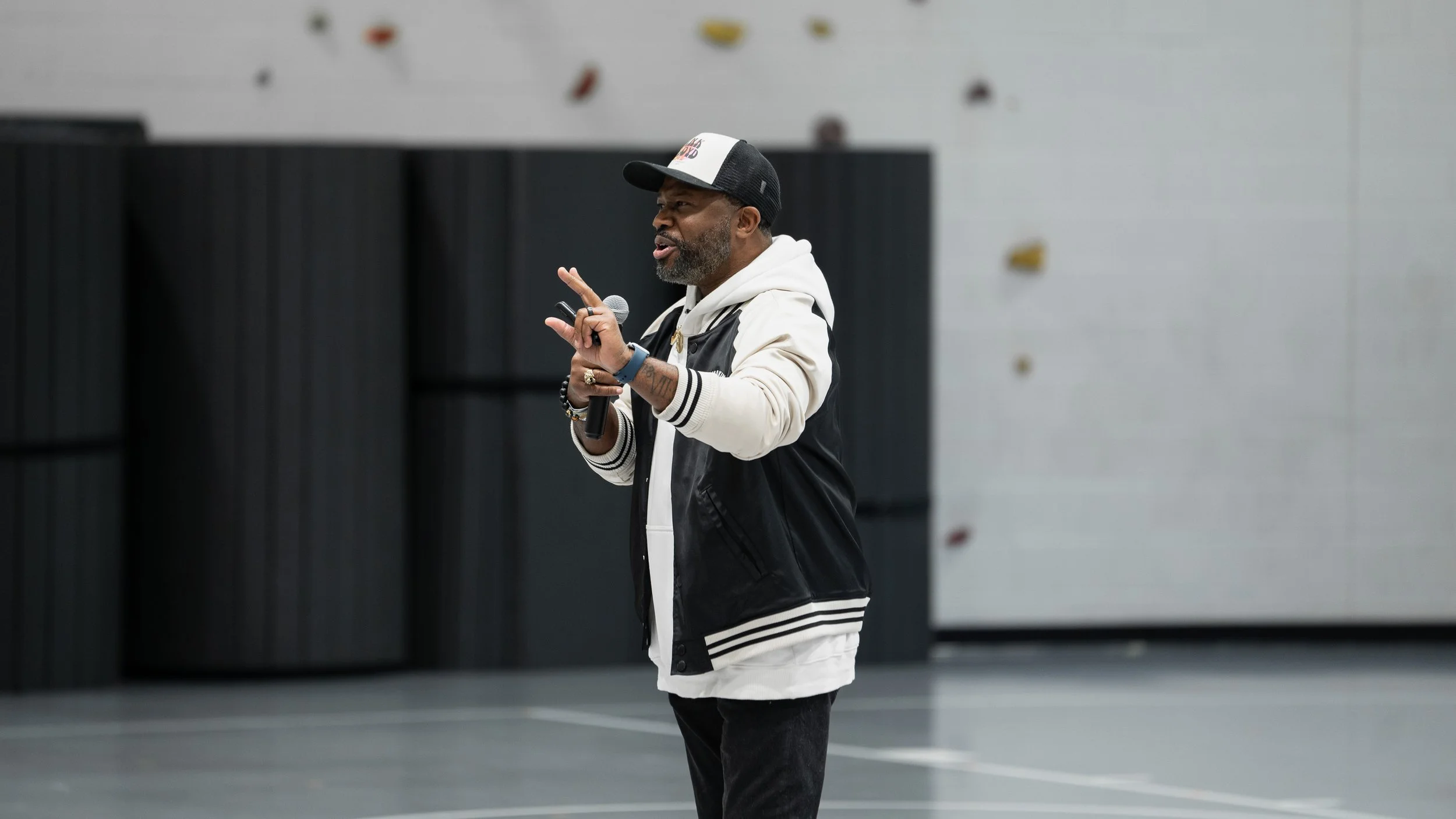 Bobby Gueh with Brothers Making Moves speaking into a microphone while gesturing with his hand in an indoor gym or climbing facility.