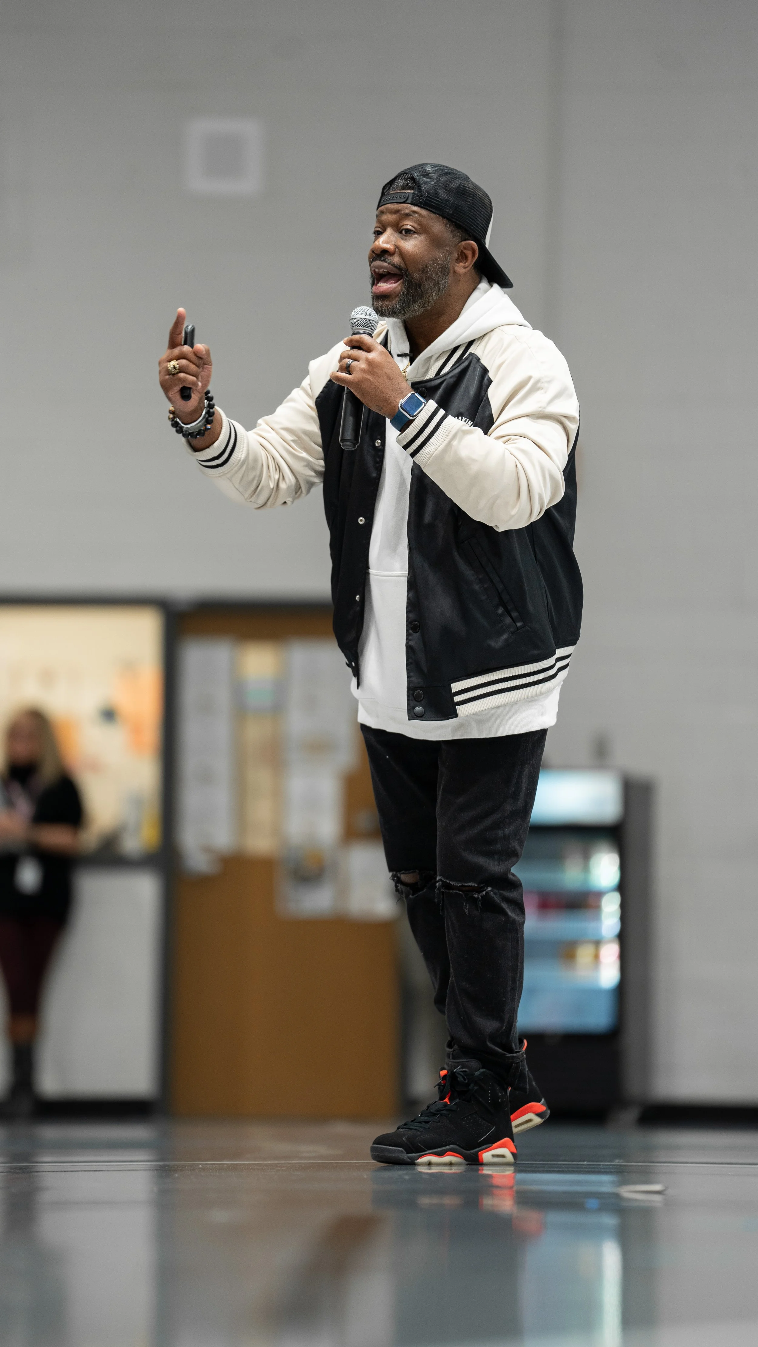 Bobby Gueh with Brothers Making Moves speaking into a microphone at an indoor event, wearing a black cap backward, a white hoodie, black distressed jeans, and black sneakers with red accents.