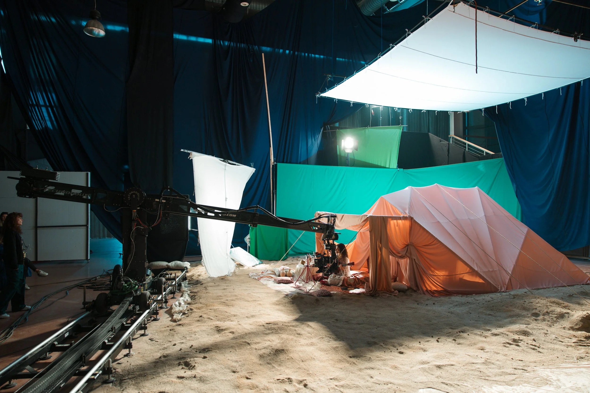 Film set with green, white, and peach-colored tents and fabric, large overhead lighting, and a sandy ground, indicating an outdoor production environment.
