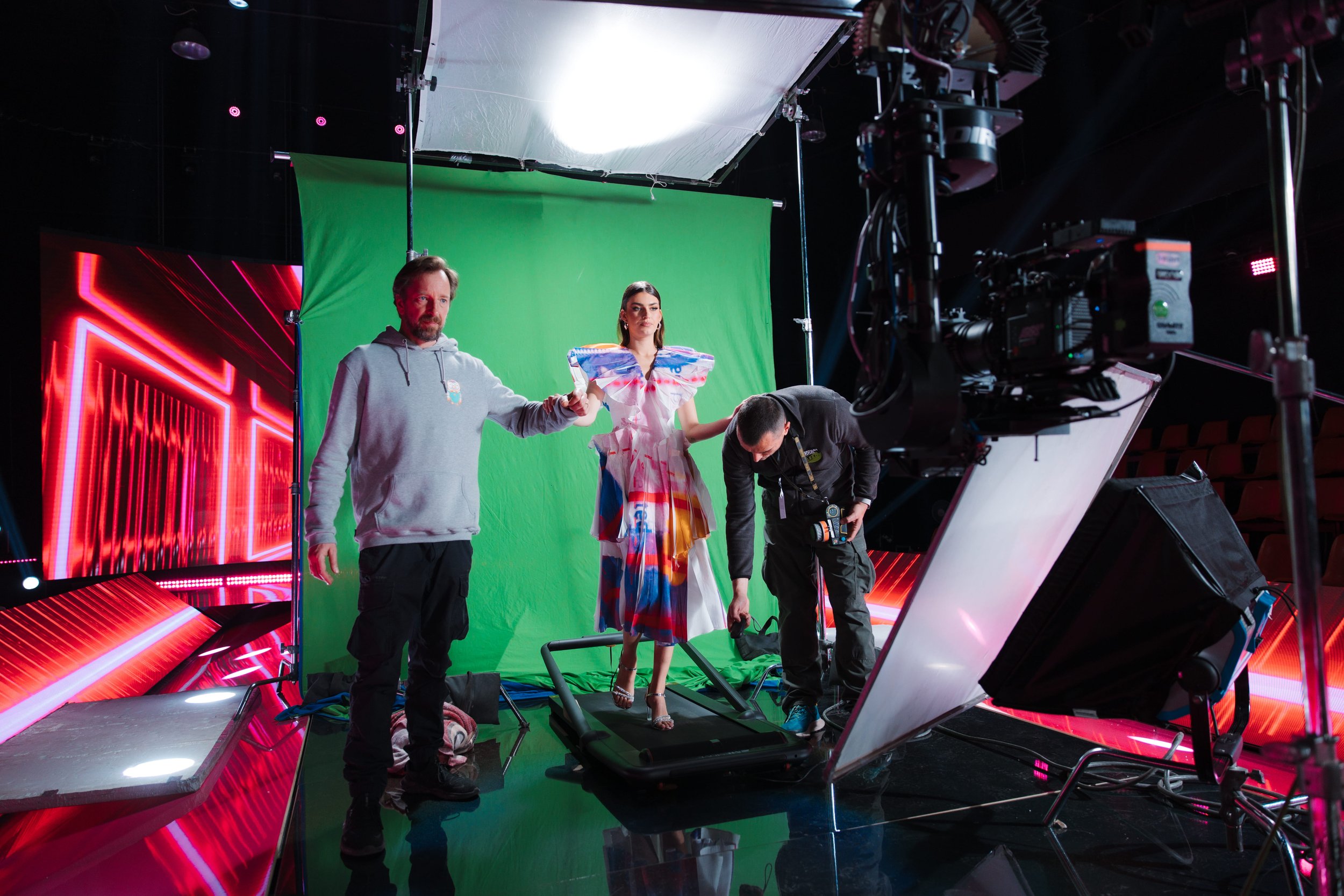Behind the scenes on a television or film set, a woman in a colorful dress stands on a treadmill, holding the hand of a man in a gray hoodie, while crew members adjust equipment and a green screen backdrop is visible behind them.