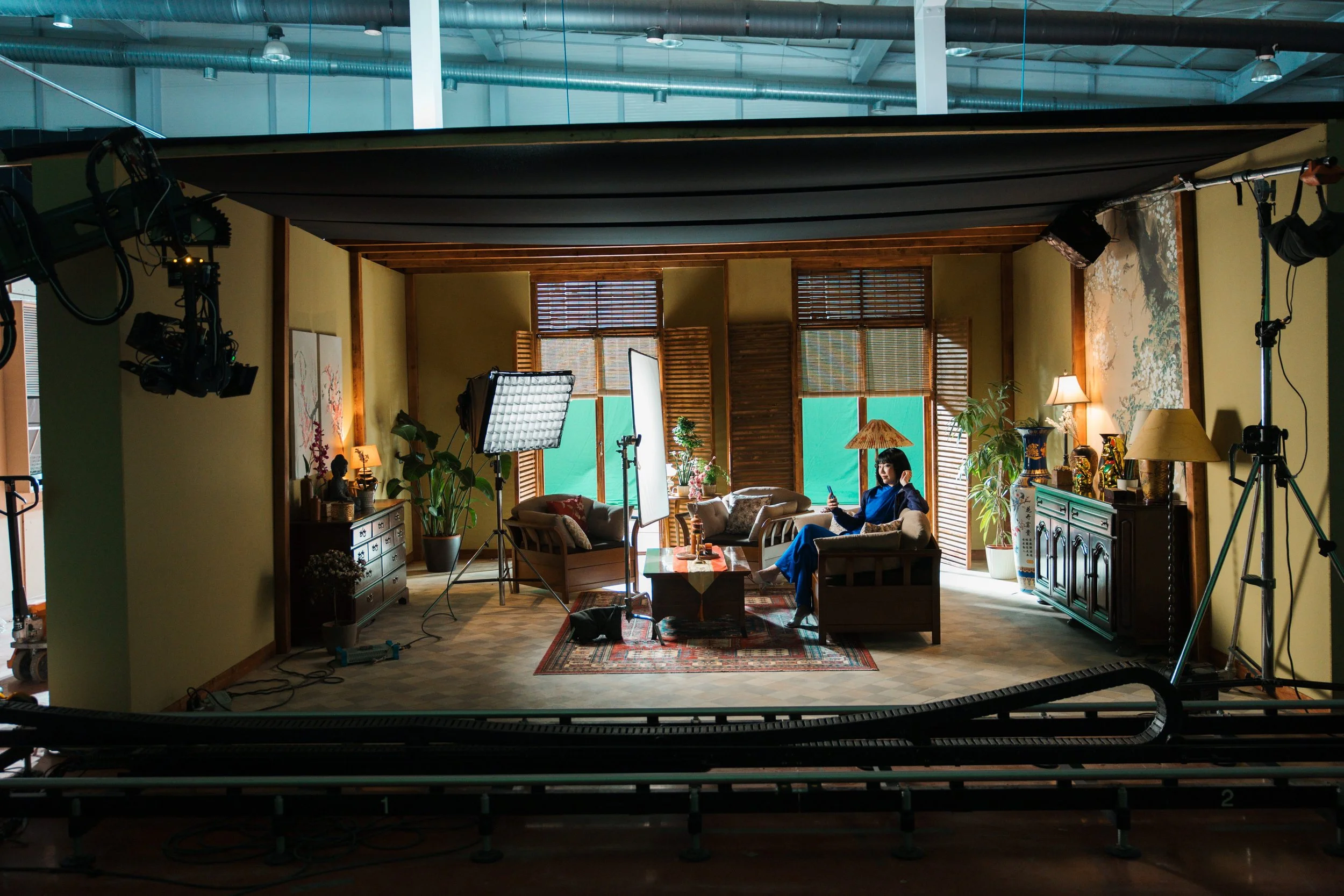 Filming set of a cozy living room with large windows, wooden blinds, plants, wall art, and a woman sitting on a sofa using her phone, surrounded by lighting and camera equipment.