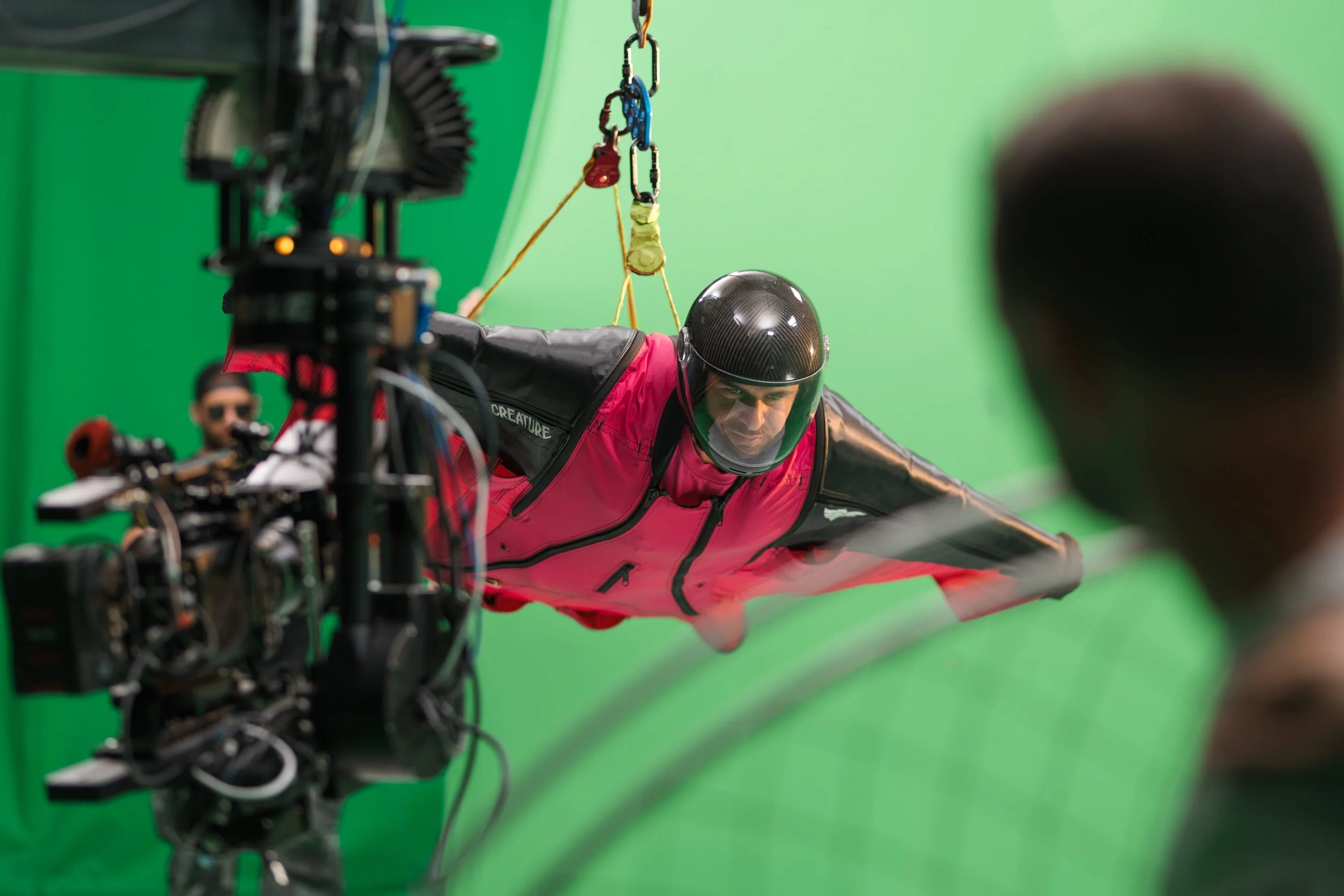 A person in a pink jacket and black helmet is lying on a green screen set, with equipment and crew members partially visible in the background, as part of a film or video production.