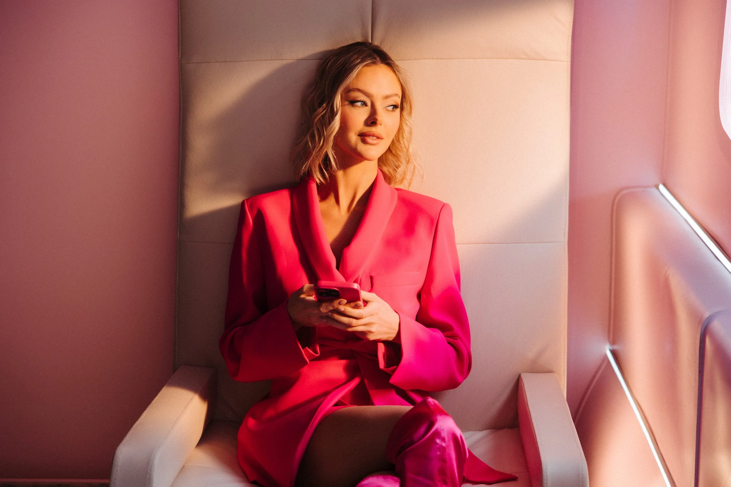 A woman in a red blazer and matching pants sitting on a pink padded seat, looking at her smartphone in a modern, softly lit space.