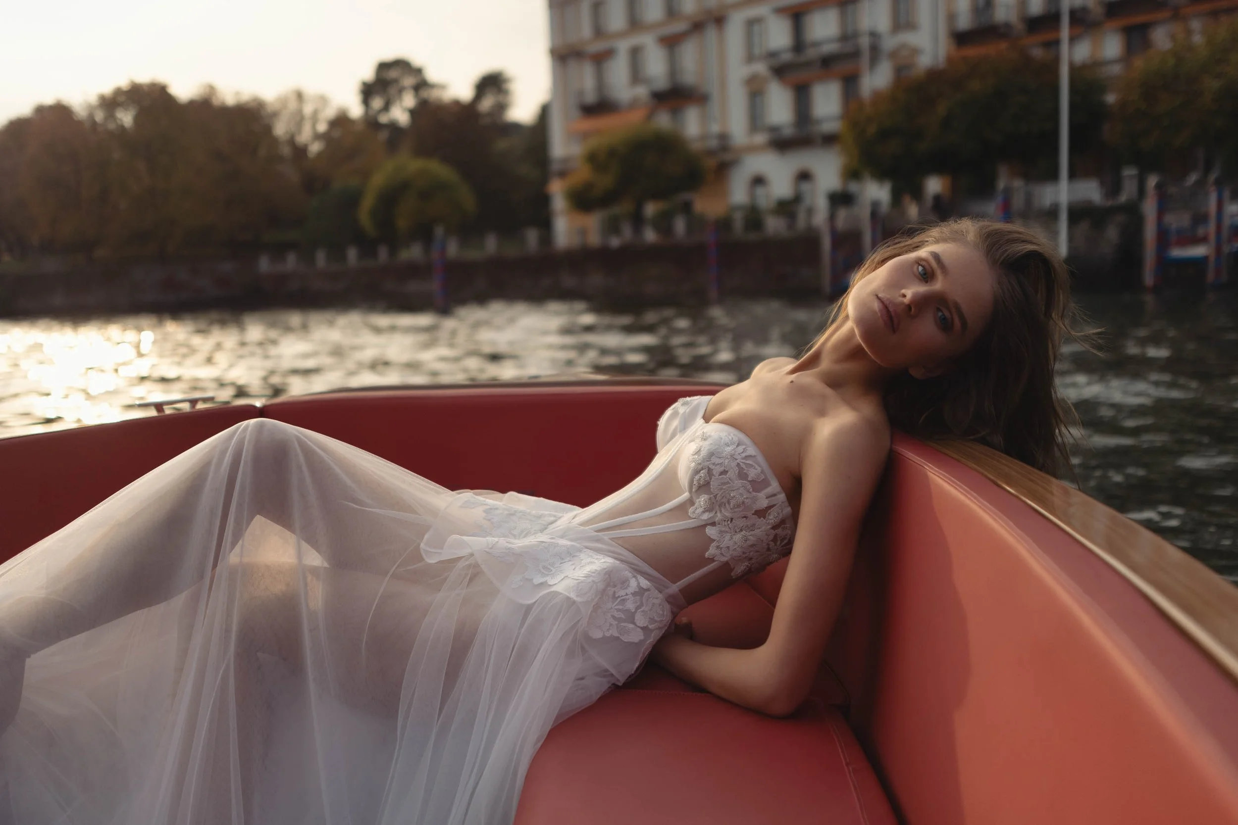 A young woman in a white lace dress lounging on a boat on a river during sunset with buildings and trees in the background.