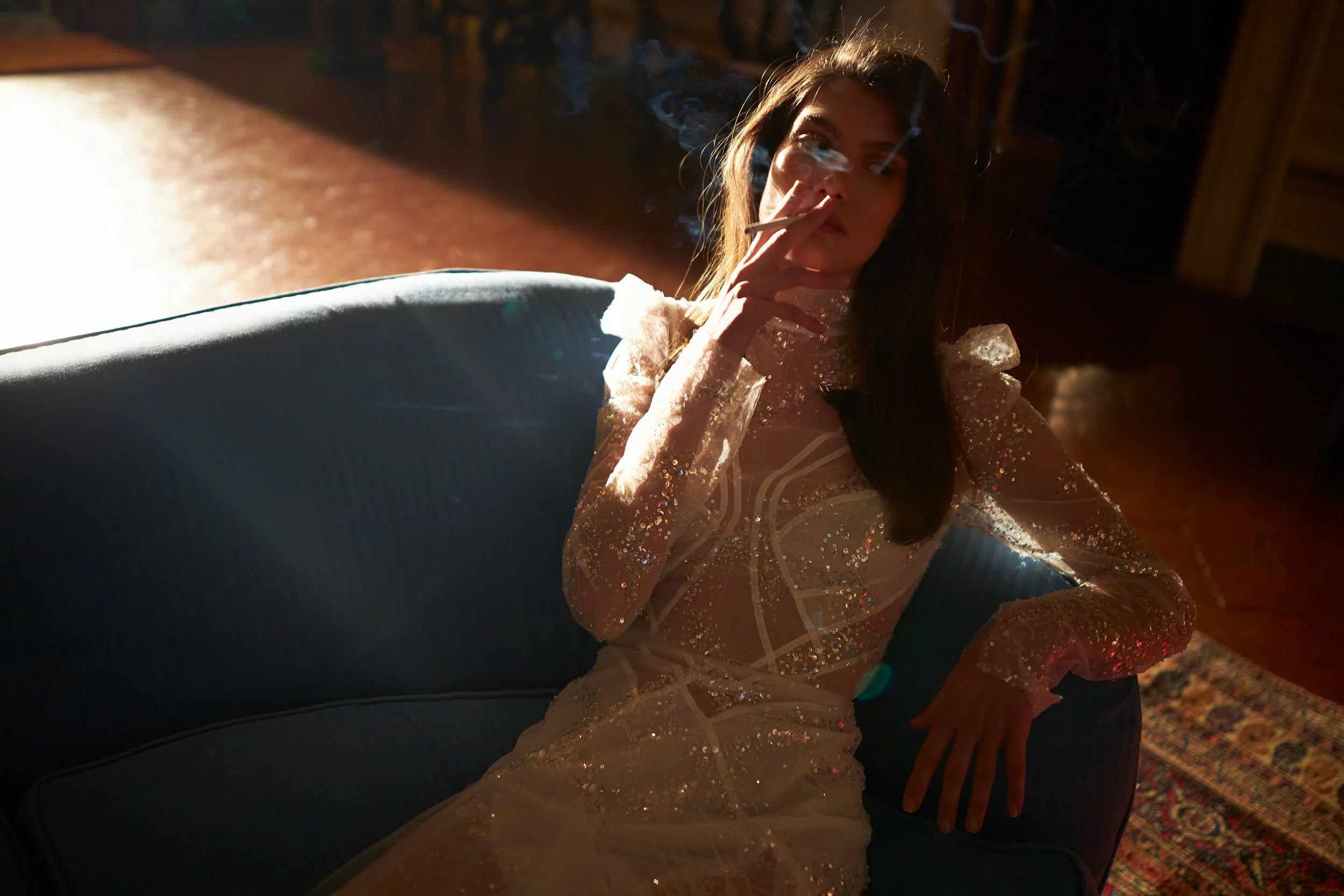 A woman in a sparkly, sheer dress sits on a blue couch in a dimly lit room, smoking a cigarette with smoke rising from her hand.