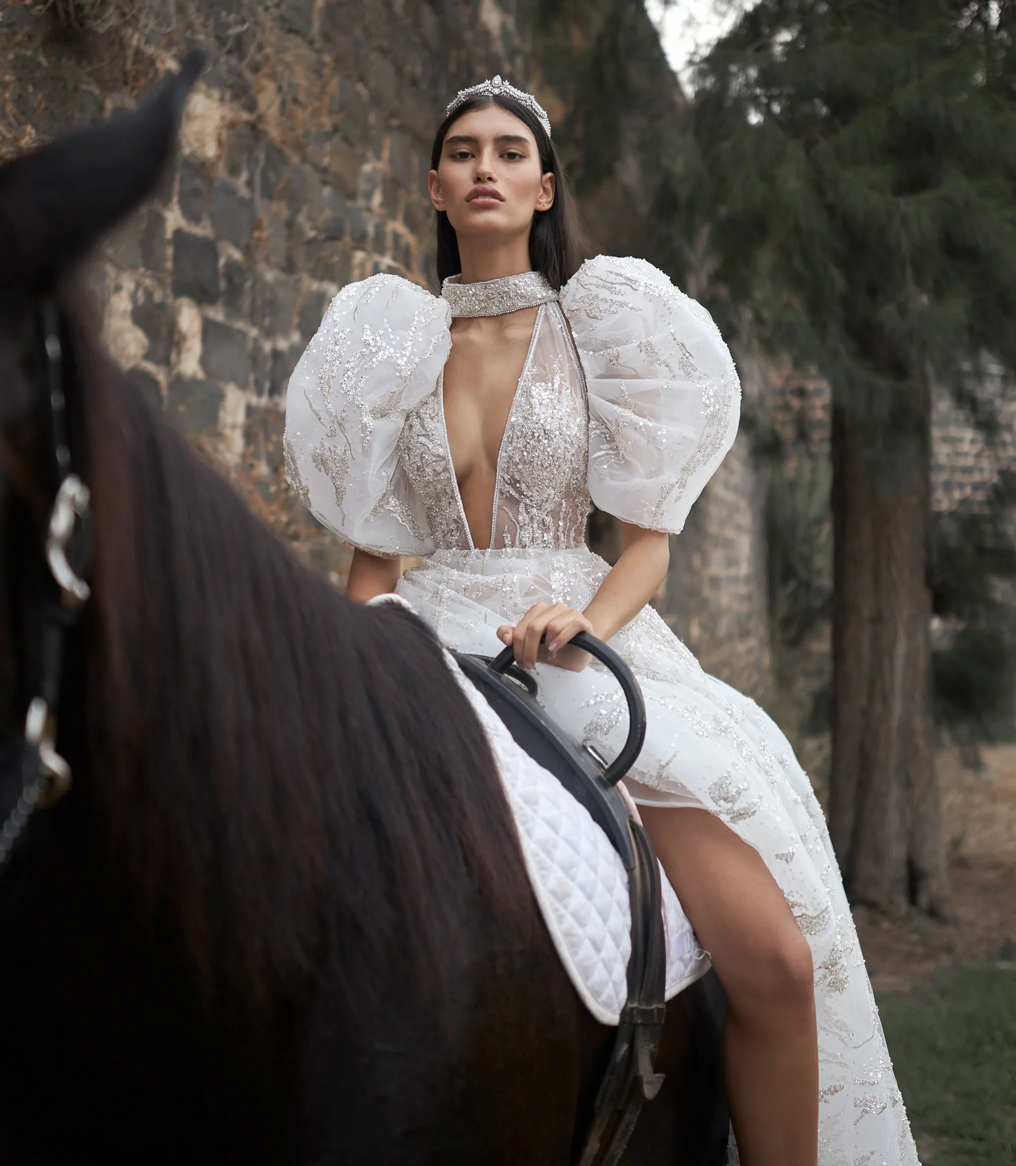 A woman in an elaborate, white, sequined dress with puffed sleeves and a tiara, riding a horse outdoors.