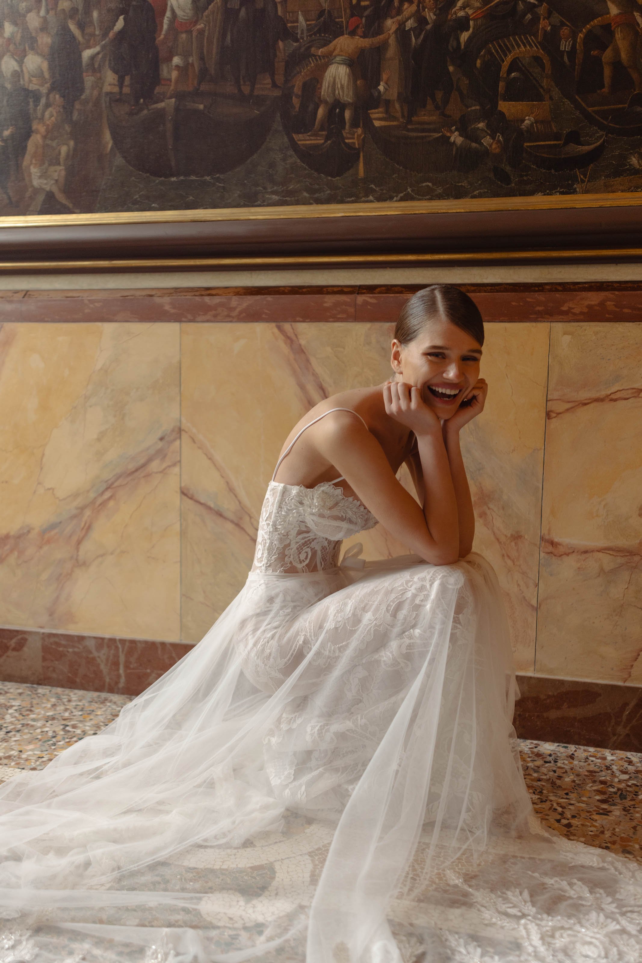 A woman in a white lace wedding dress is sitting on the floor, smiling with her hands resting on her cheeks, against a marble wall with a mural above.