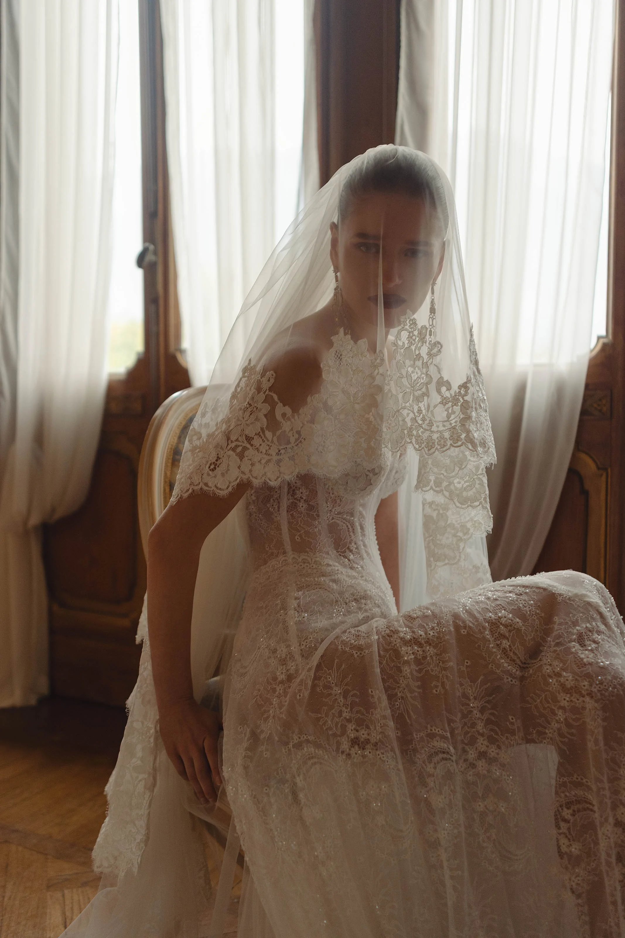 A woman in a lace wedding dress and veil sitting in a room with wooden furniture and cream curtains, looking directly at the camera.