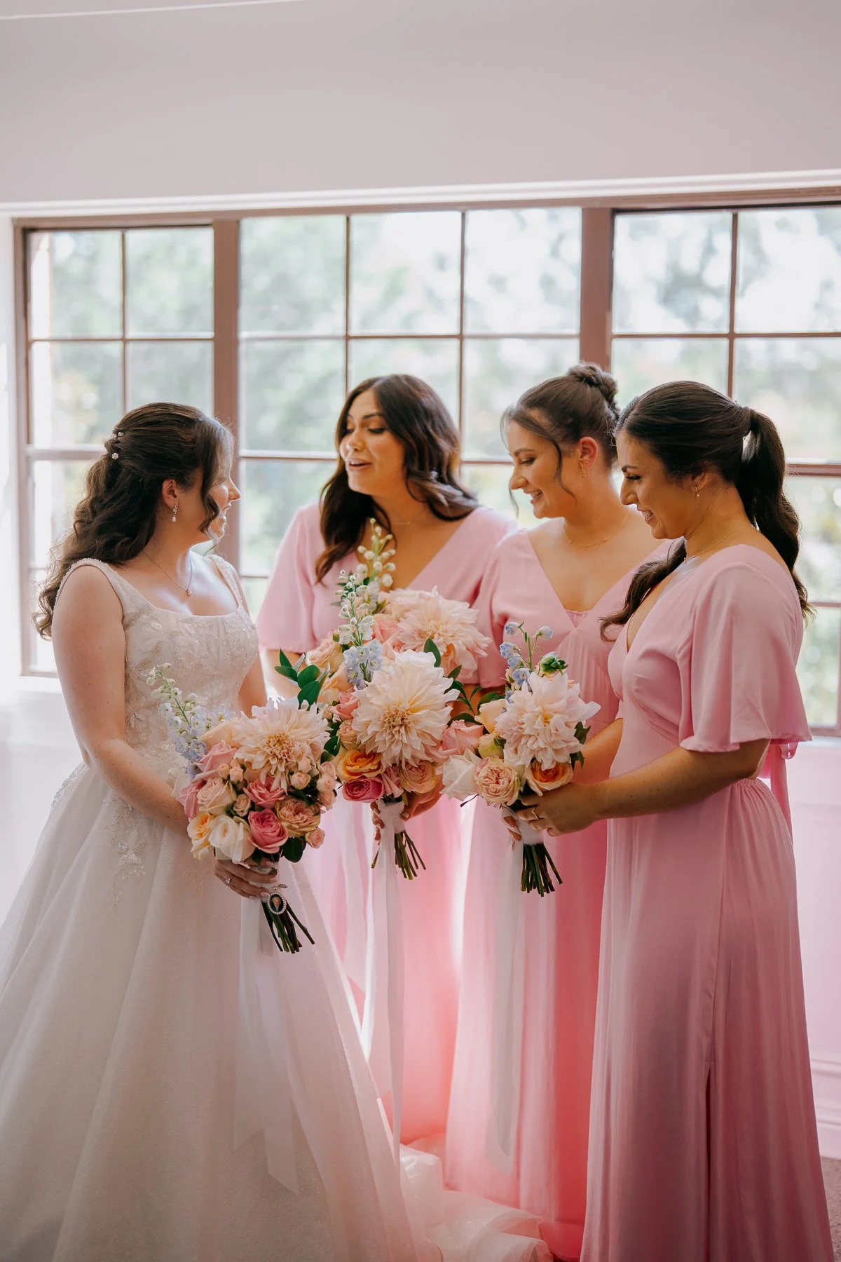 A bride with brown hair in a white wedding gown holding a bouquet of pink and white flowers, standing with four women dressed in pink, all holding similar bouquets, inside a room with large windows.