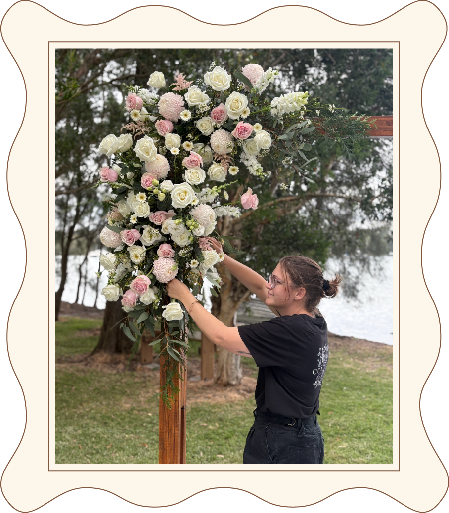 Person arranging a large floral display with white and pink roses, white dahlias, and greenery outdoors near a body of water.