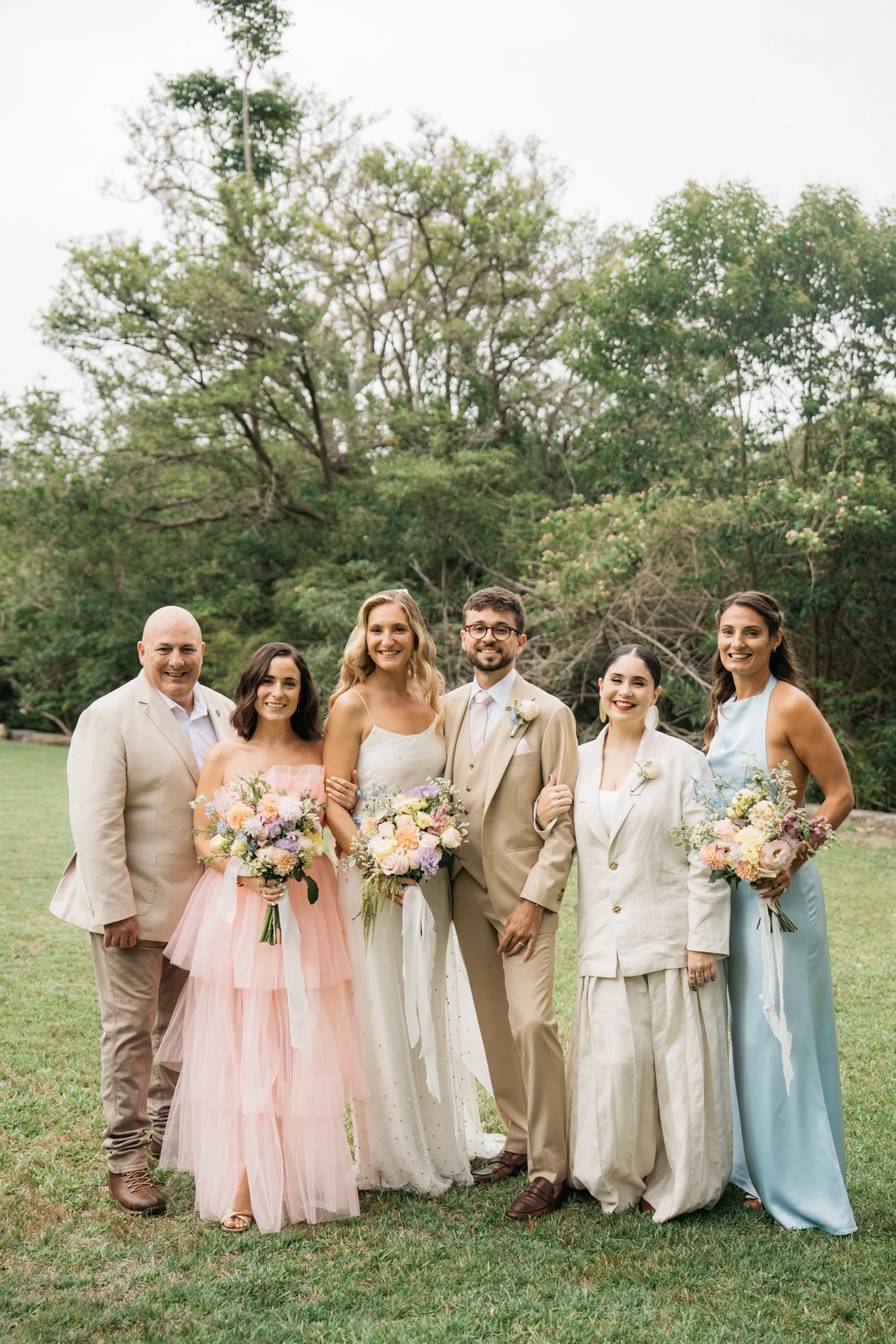 A group of seven people in formal attire standing outdoors on grass with trees in the background, celebrating a wedding with some holding flower bouquets.
