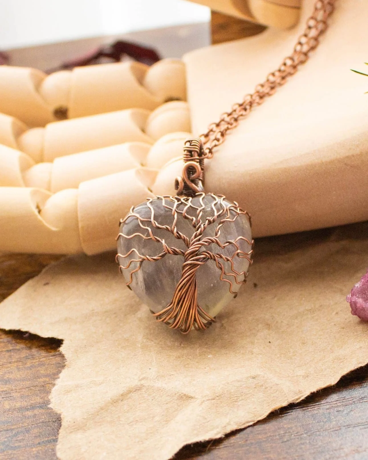 A heart shaped pendant wrapped in copper wire designed like a tree, with a copper chain, resting on a piece of paper on a wooden hand model.