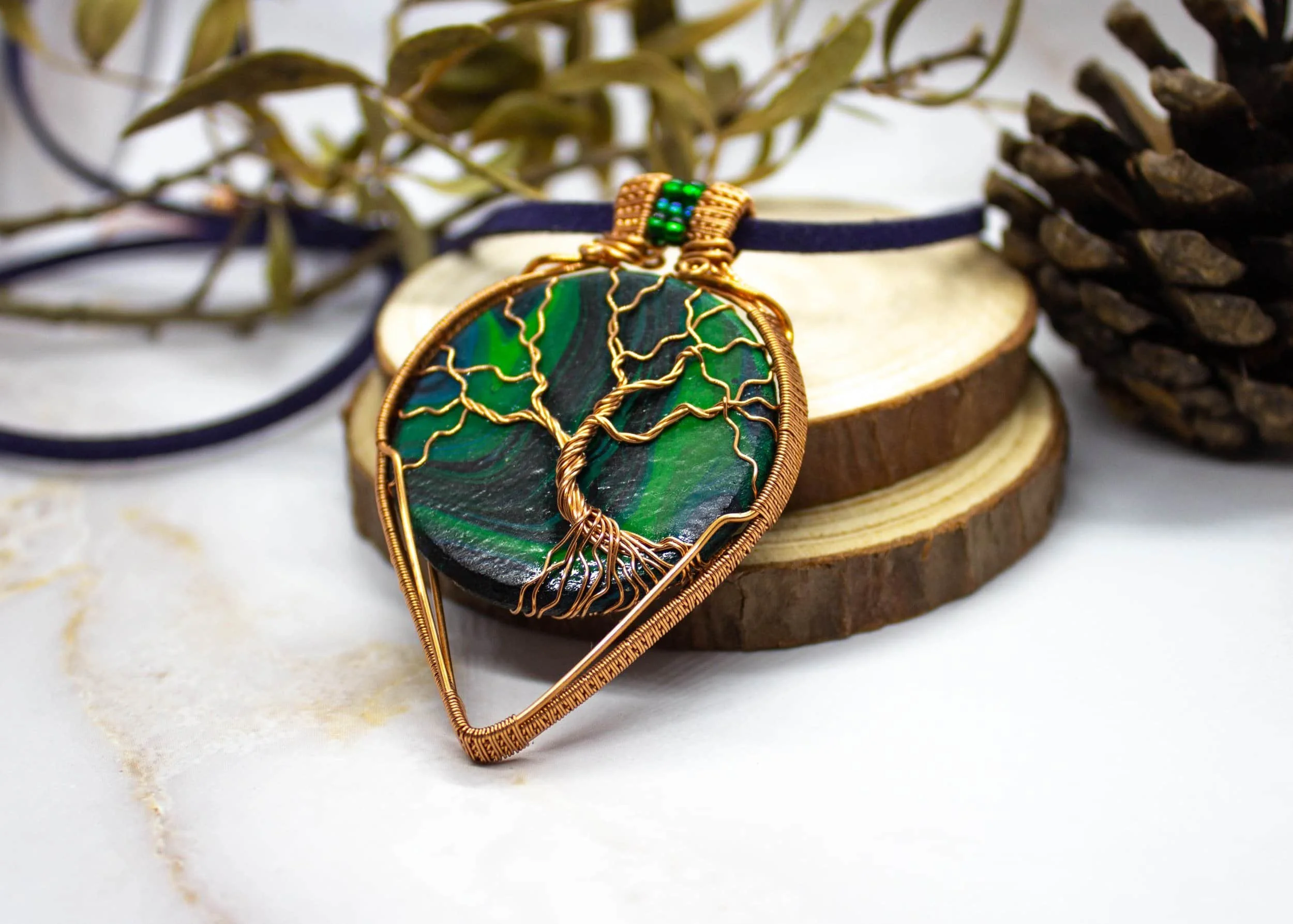 A pendant with a tree of life design made of wire wrapping a hand painted wood piece, resting on a wooden slice with pinecones and leaves in the background.
