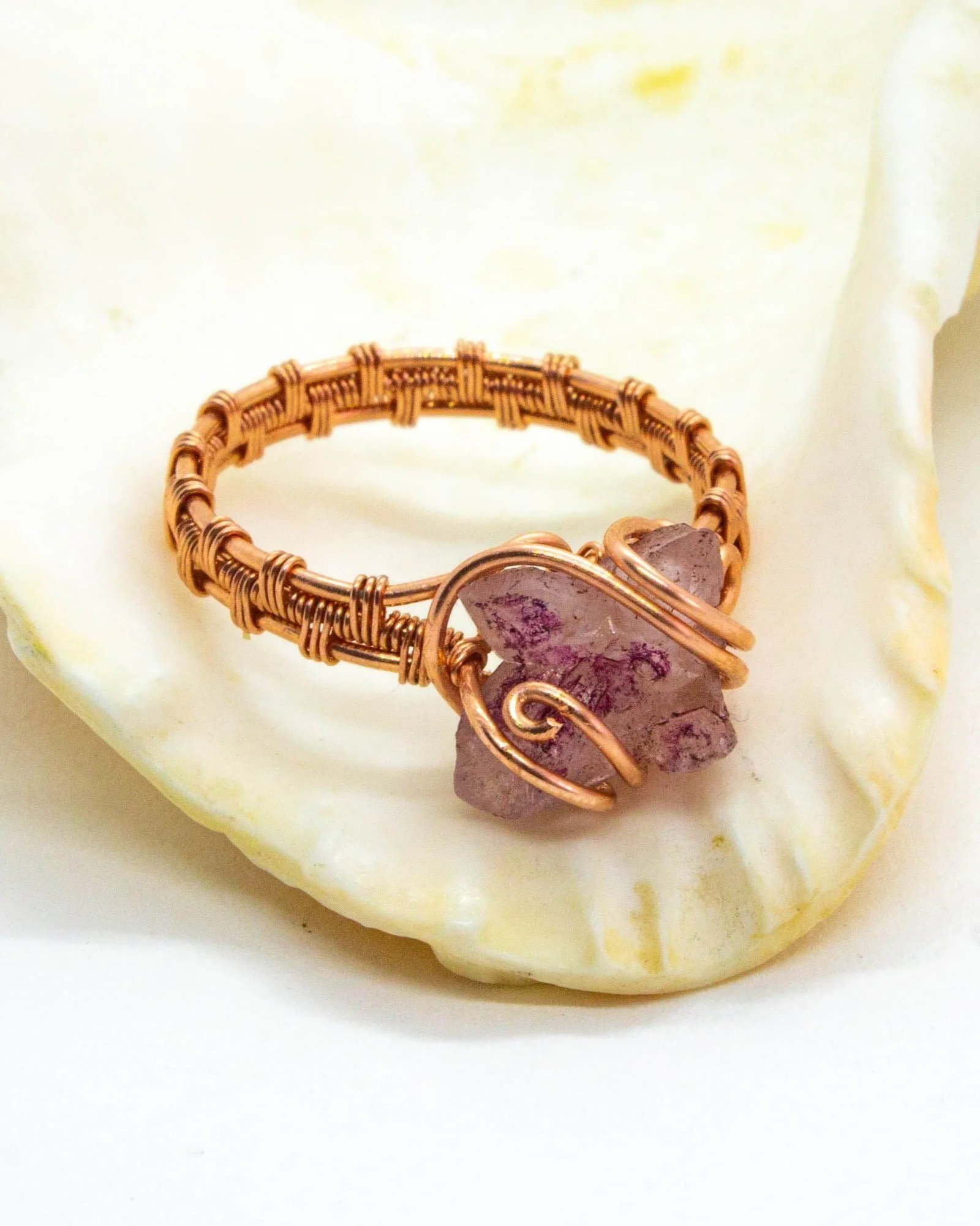 A copper wire wrapped ring with a rose quartz stone centerpiece, resting on a large white shell.