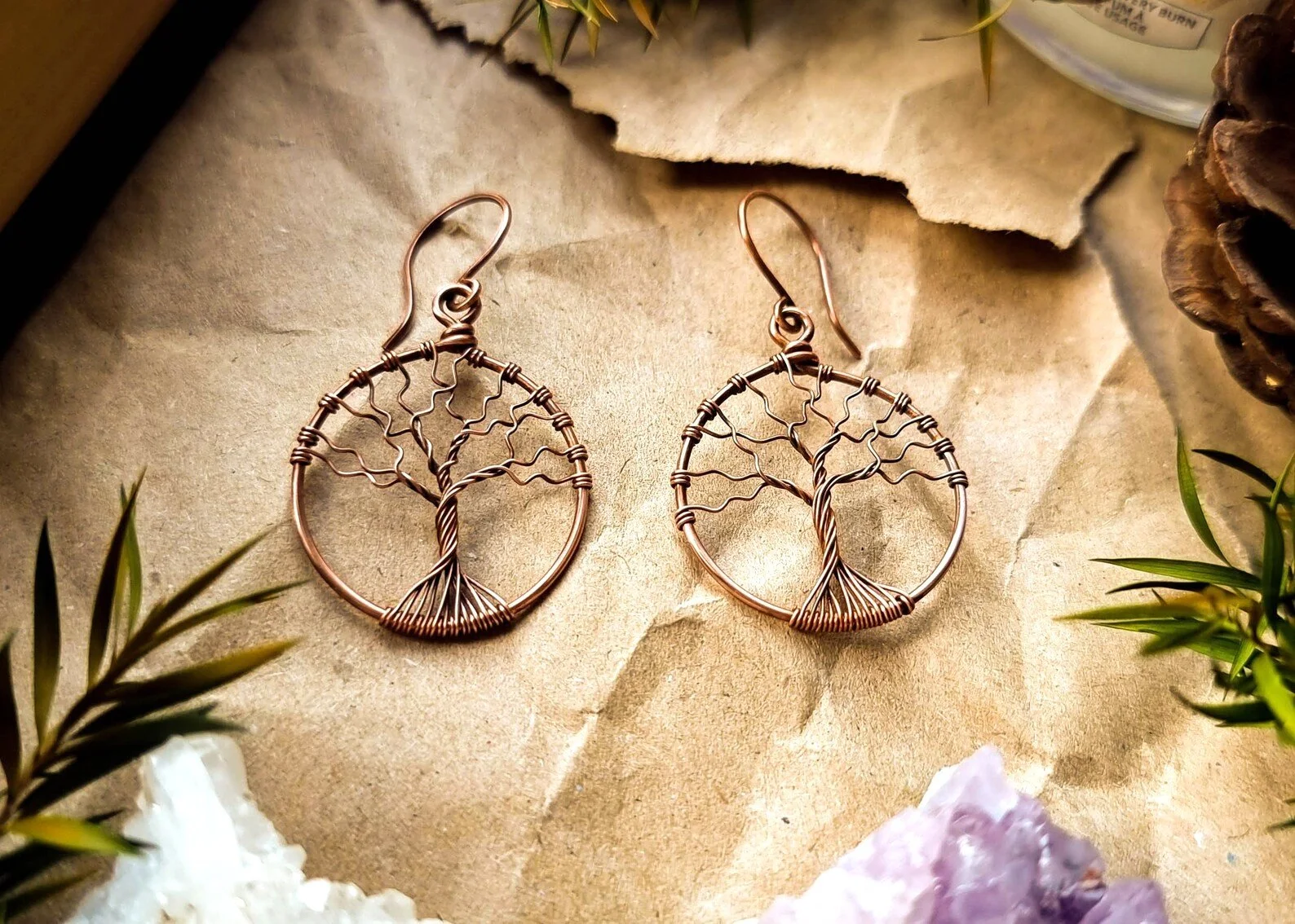 Copper wire tree of life earrings on brown card, surrounded by crystals and greenery.