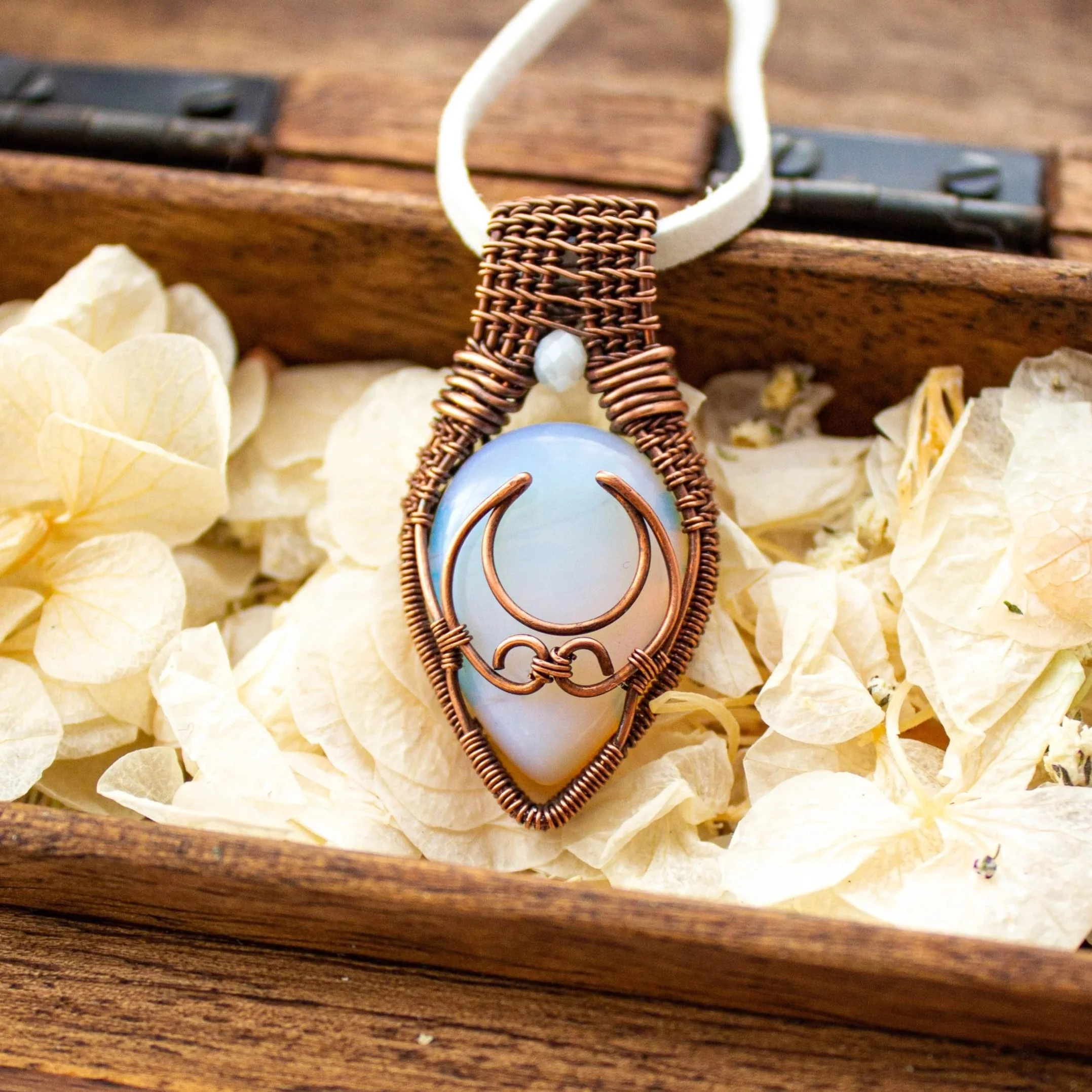 Placed in a wooden box with flower petals is a wire wrapped copper pendant with a white stone and a crescent moon copper accent