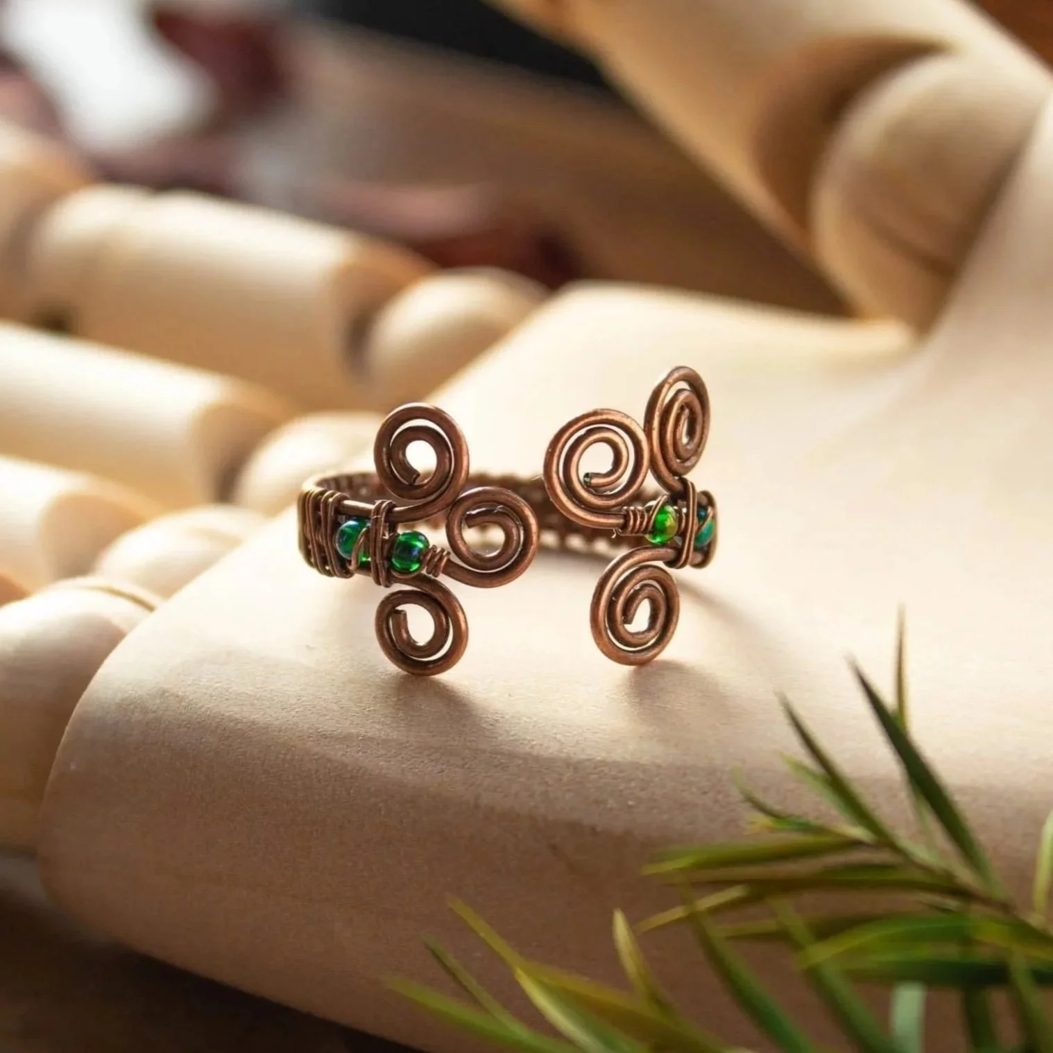 A copper wire ring with green beads, designed with multiple spiral patterns, placed on a wooden model hand, surrounded by leaves.