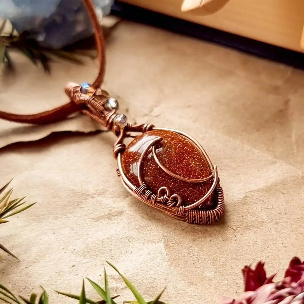 Handcrafted pendant with a copper wire wrapped around a glittering brown stone, placed on a textured surface surrounded by greenery and crystals.