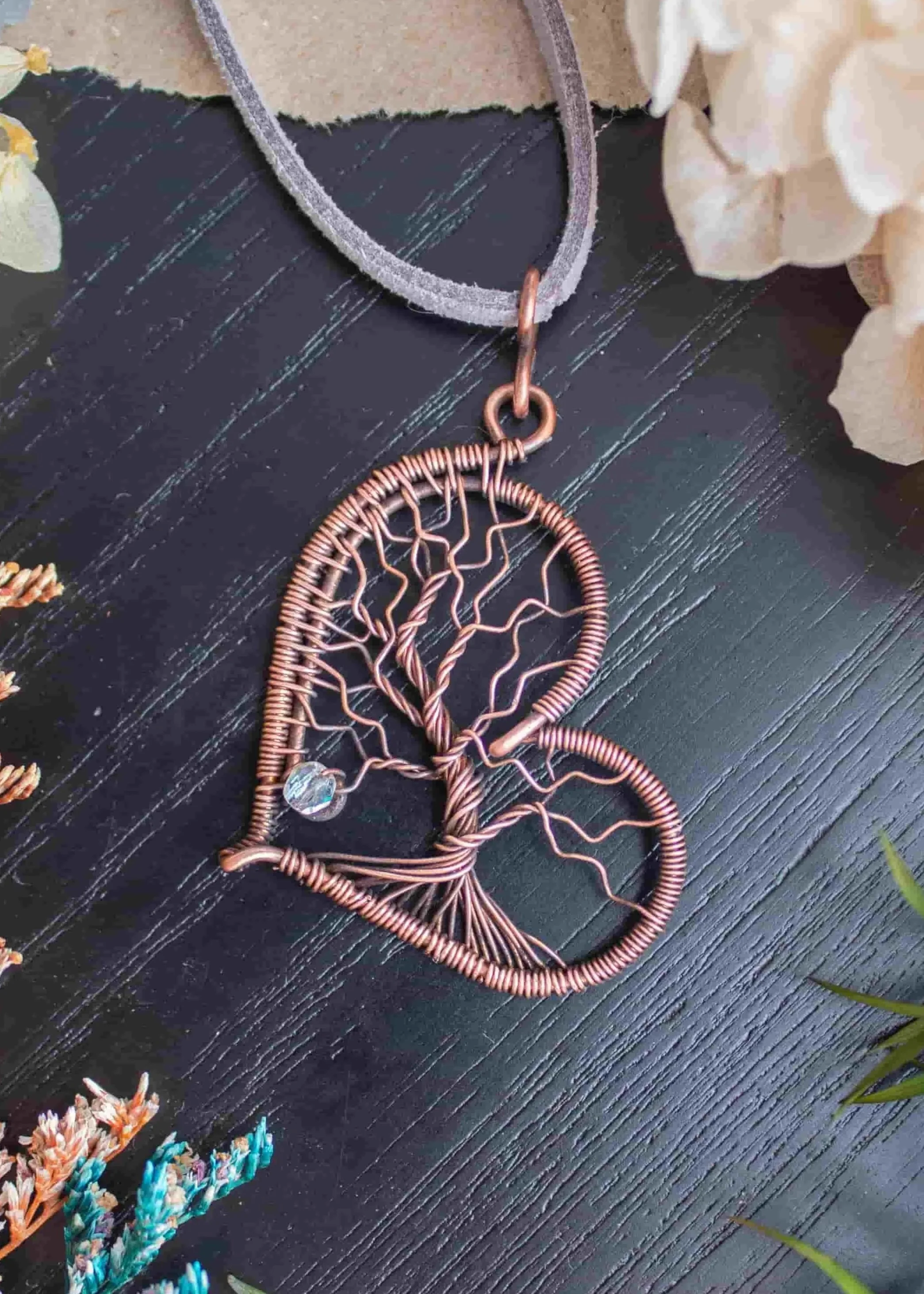Resting against a black wood background is a wire wrapped copper pendant in the shape of a heart with a tree of life inside it with a glass bead accent on one of the branches.