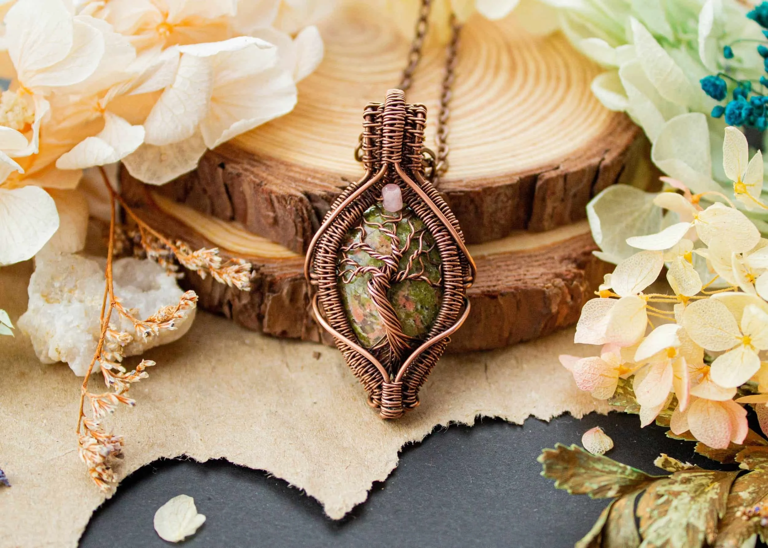Wire-wrapped pendant featuring a tree design encasing a unakite stone, surrounded by dried flowers and placed on a piece of wood.