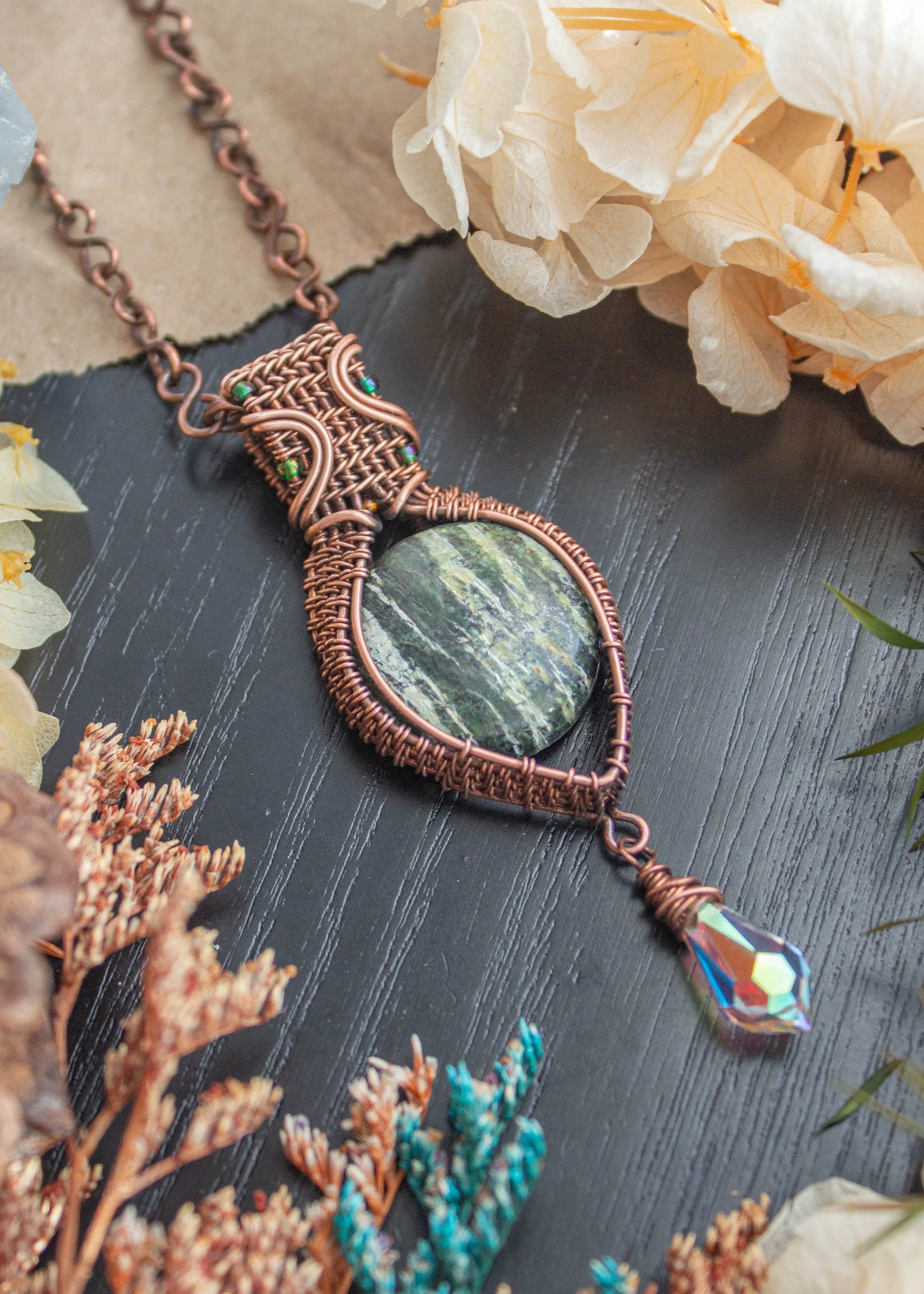 Resting against a black wood background is a wire wrapped copper pendant with a round green stone centrepiece and a crystal briolette attached to the bottom.