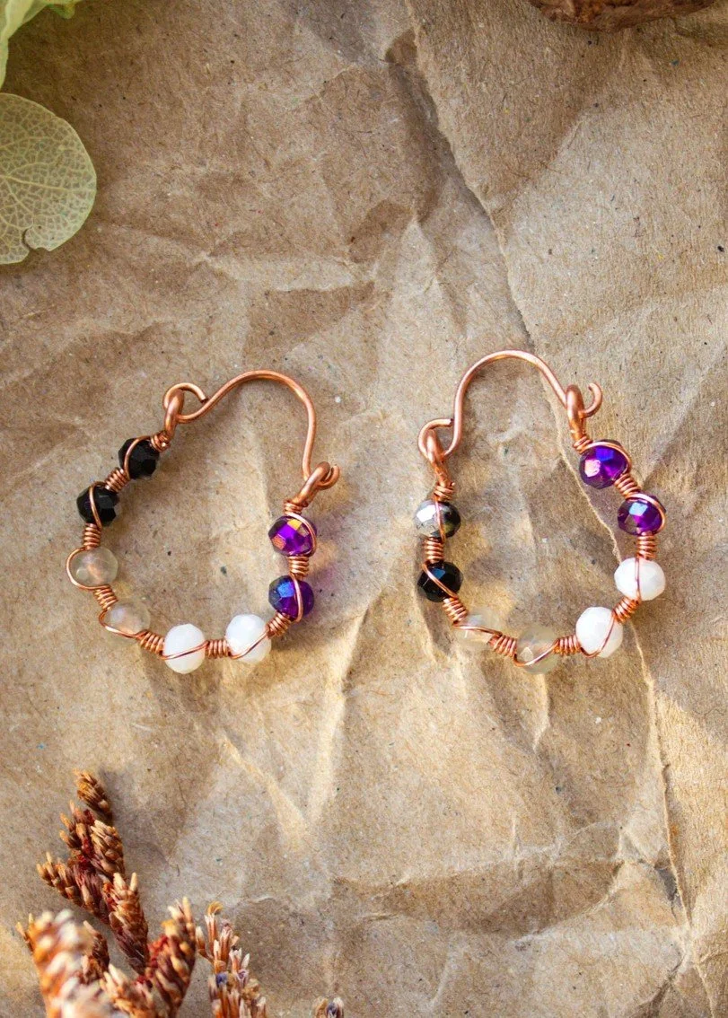 A pair of handmade wire earrings with beads arranged by the colours of the asexual flag, displayed on a textured brown paper background.