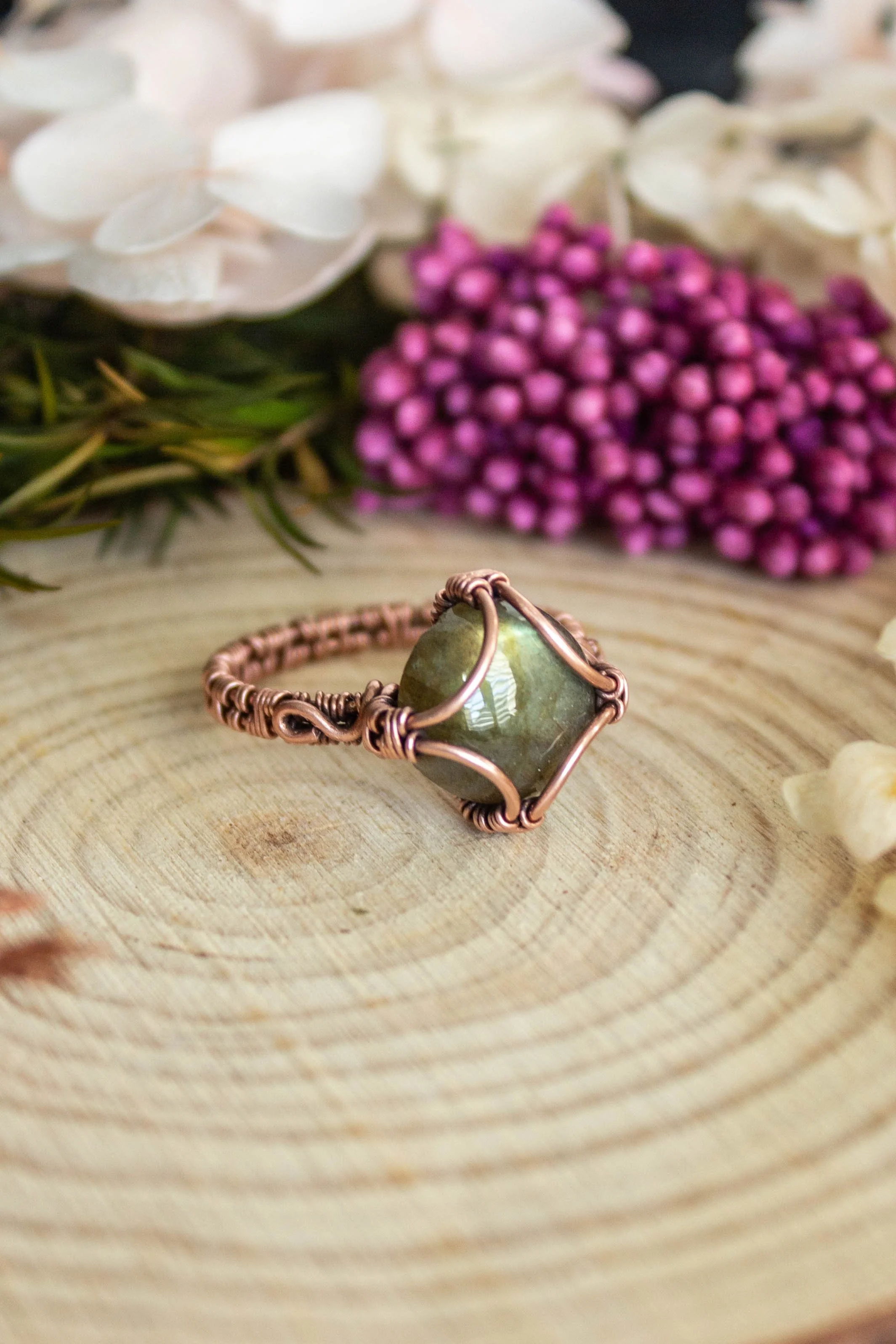 A wire-wrapped ring with a green labradorite stone on a wooden surface, with a background of white, purple, and pink flowers.