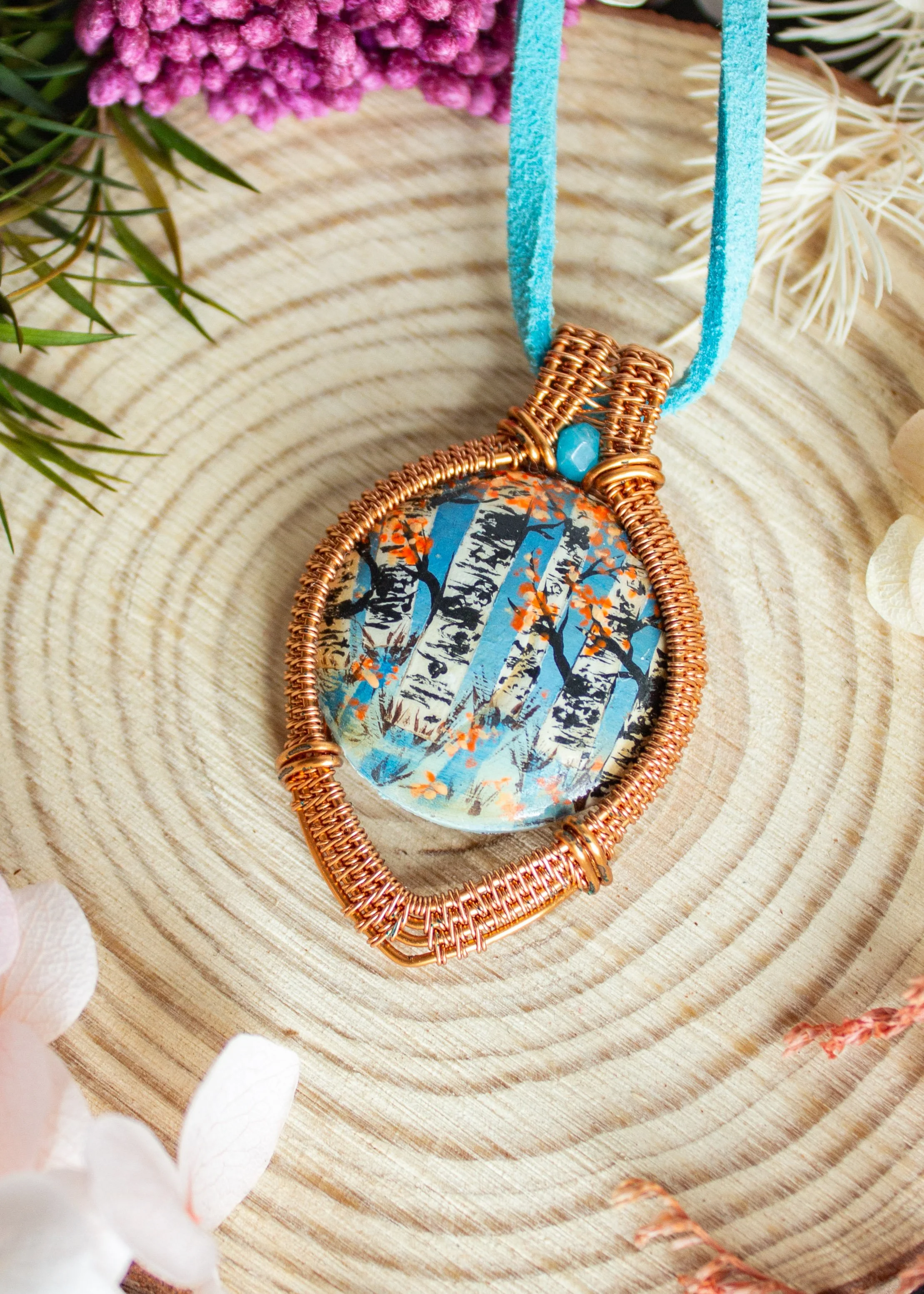 A pendant necklace with a colorful, abstract design featuring trees and orange leaves, wrapped in copper wire and hanging from a light blue cord, placed on a wooden surface surrounded by flowers and artificial plants.