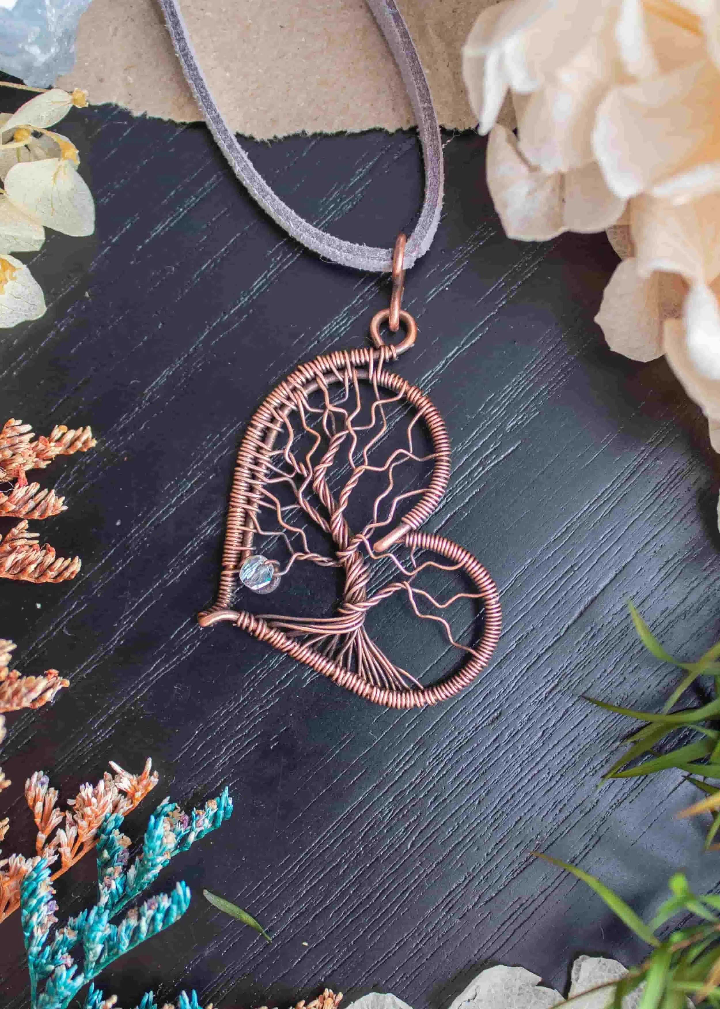 Heart shaped pendant with an inlaid tree of life deisign, resting on a black wood surface and surrounded by leaves and flowers.