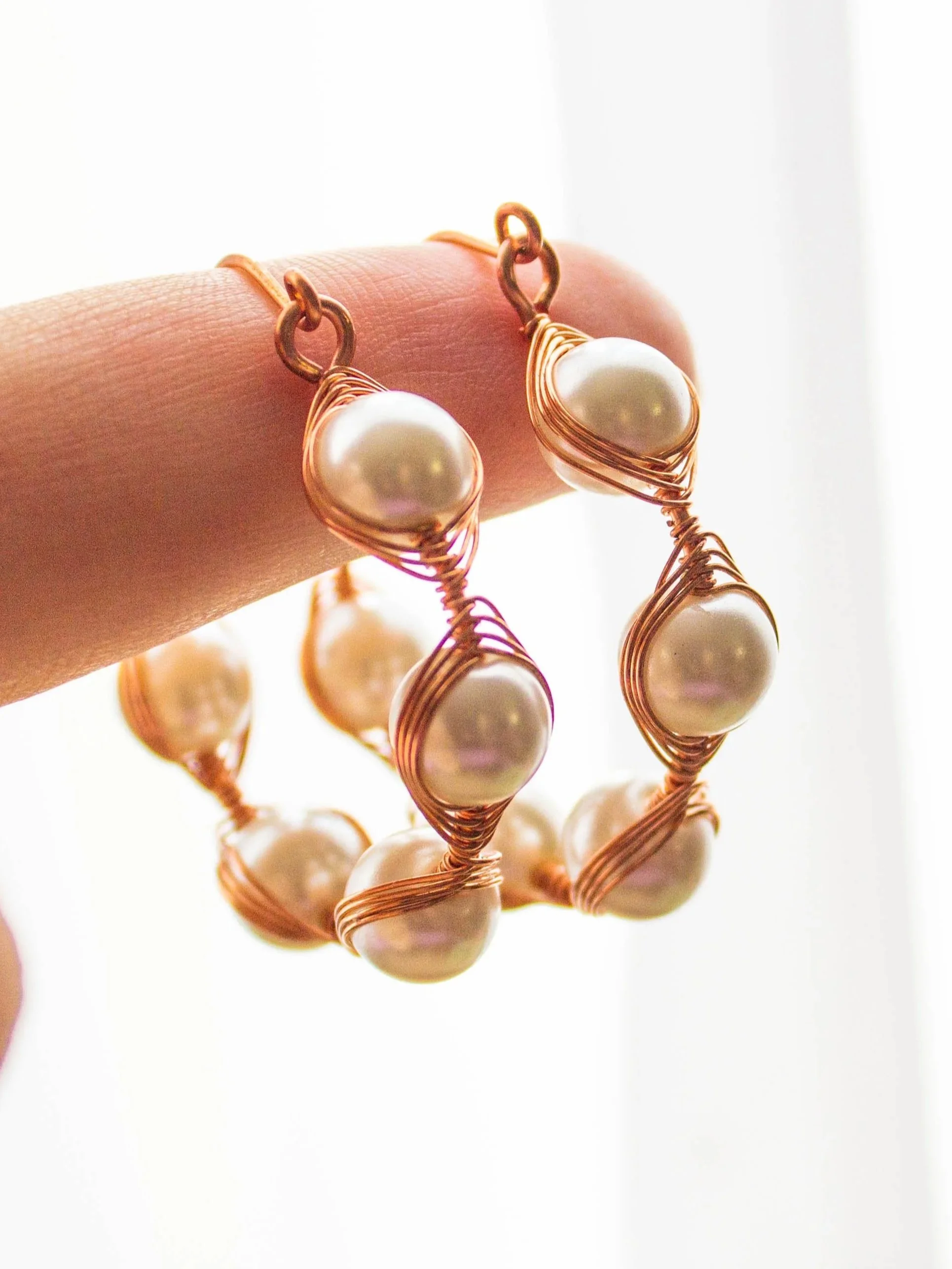 Dangling from a finger against a white background a pair of wire wrapped copper hoops with white beads