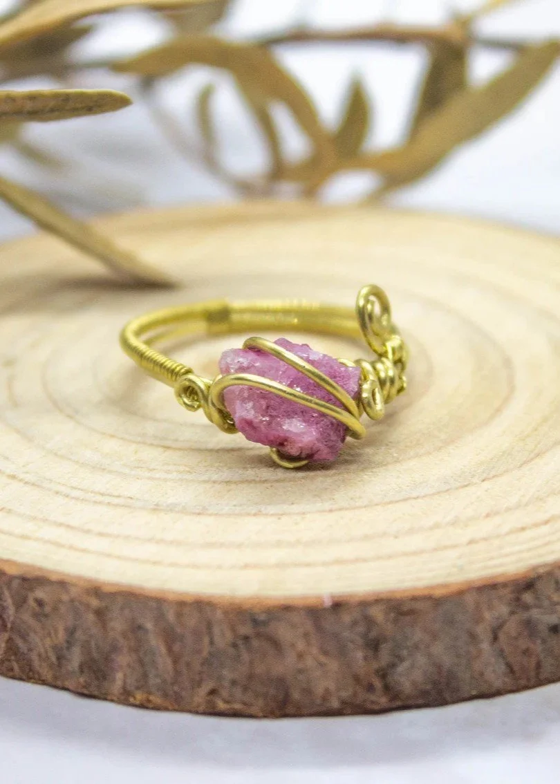 A brass wire wrapped ring with a pink stone at its center, displayed on a wooden surface.