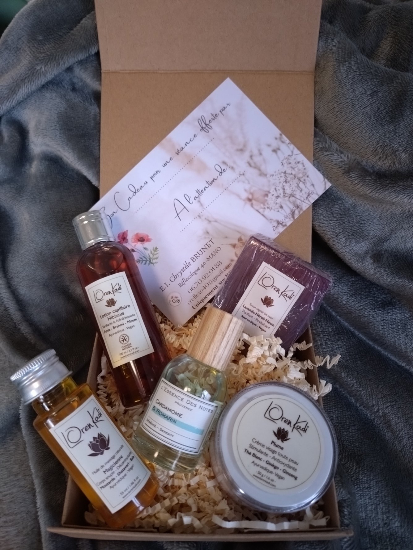 Coffret cadeau contenant plusieurs produits de soins naturels tels qu'une lotion capillaire, un parfum aux huiles essentielles, une crème pour la peau et un savon, présenté dans un coffret avec un bon pour une séance de réflexologie.