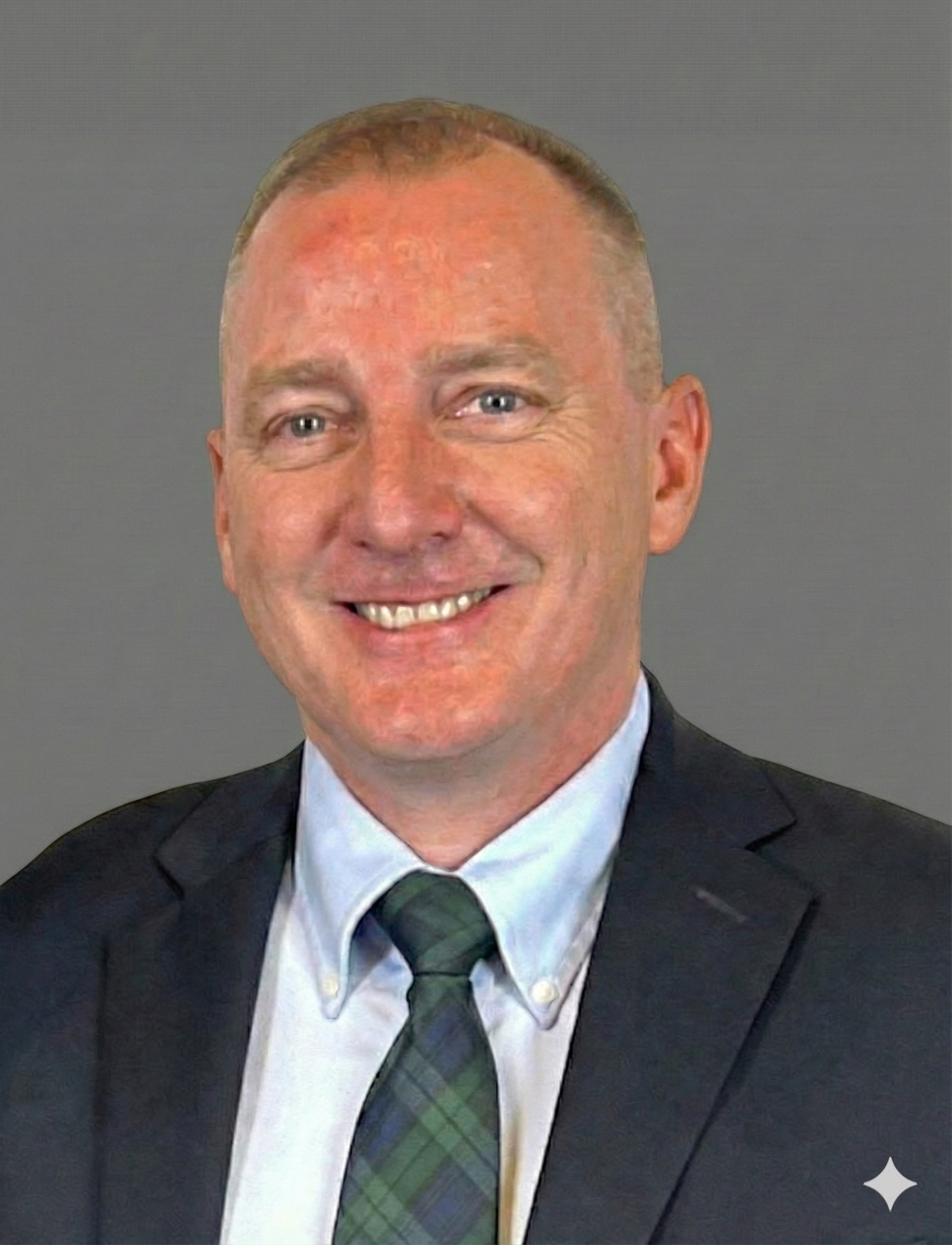 A portrait of a smiling man in a suit with a green plaid tie, against a gray background.