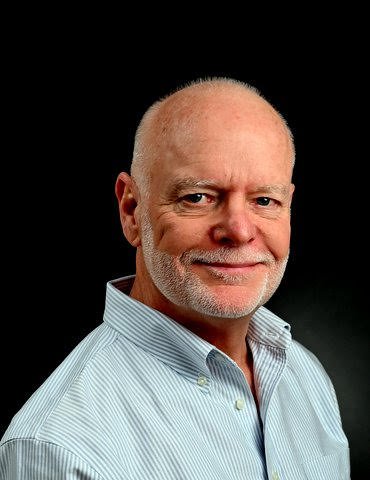 Portrait of an older man with a white beard and mustache, wearing a light blue button-up shirt against a black background.