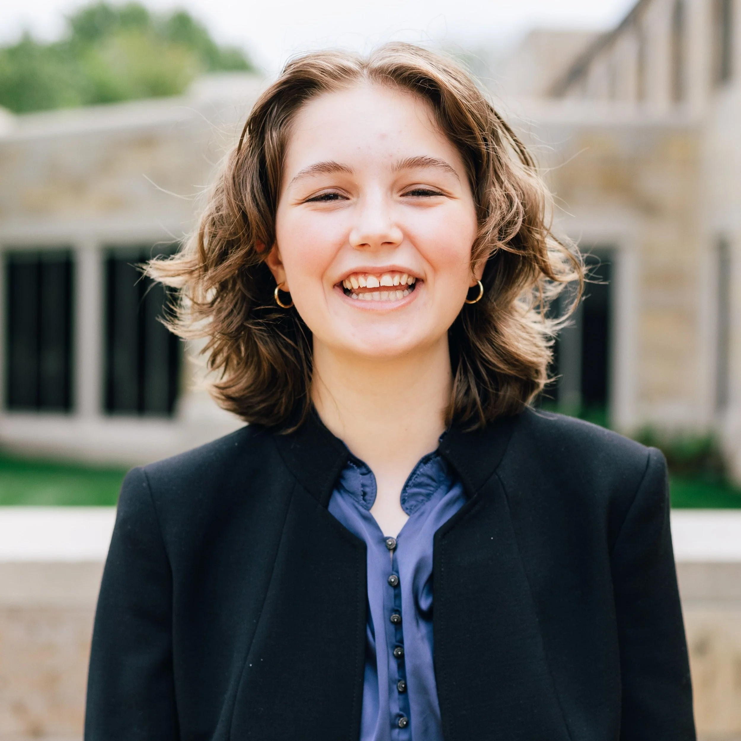 A young woman with short, wavy brown hair, wearing a black blazer and a dark blue blouse, smiling outdoors in front of a blurred building and greenery.