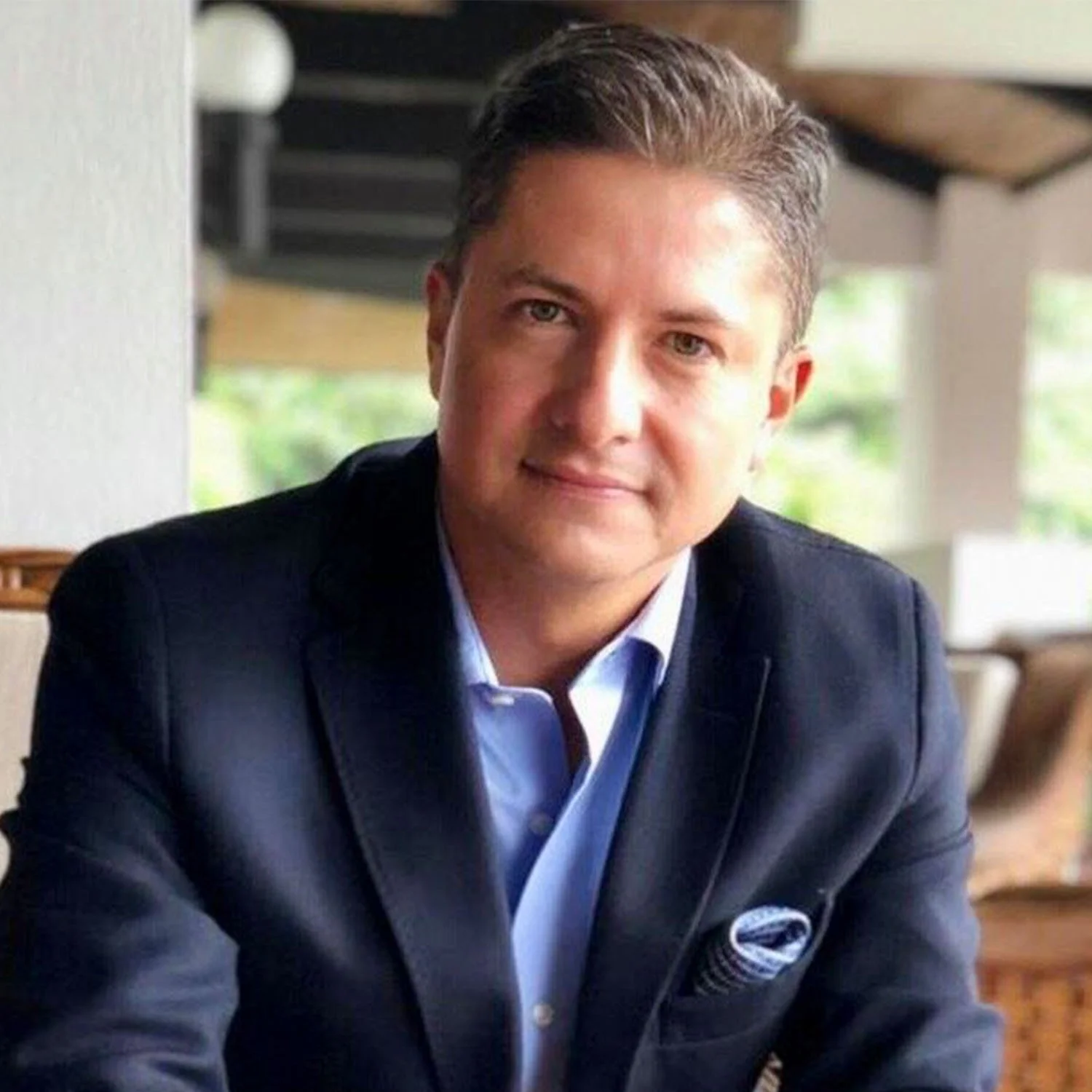 A man wearing a navy blazer and light blue shirt sitting indoors with a blurred background of greenery and furniture.