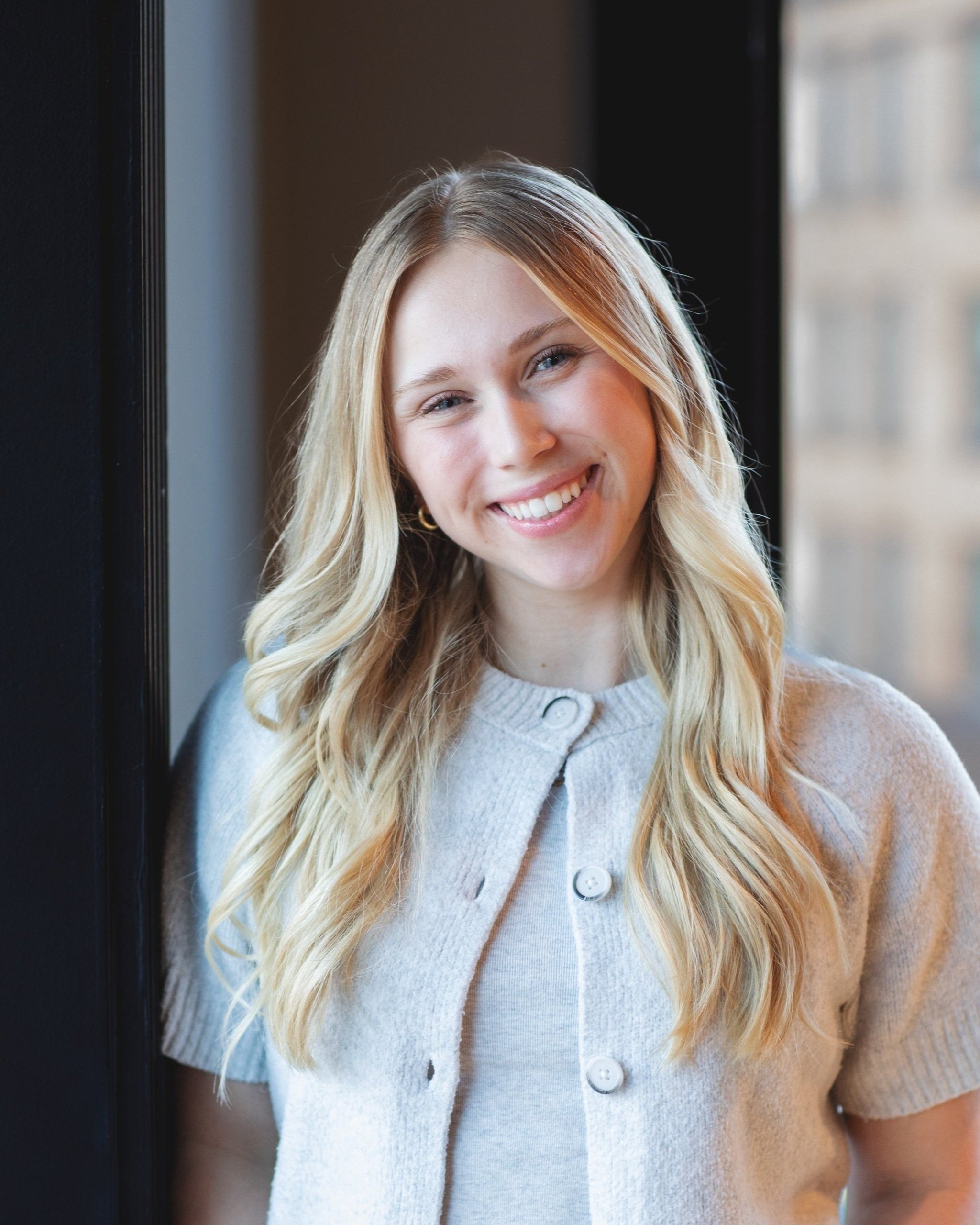 Young woman with long wavy blonde hair smiling, wearing a light blue shirt and a gold heart necklace, standing against a plain beige wall.