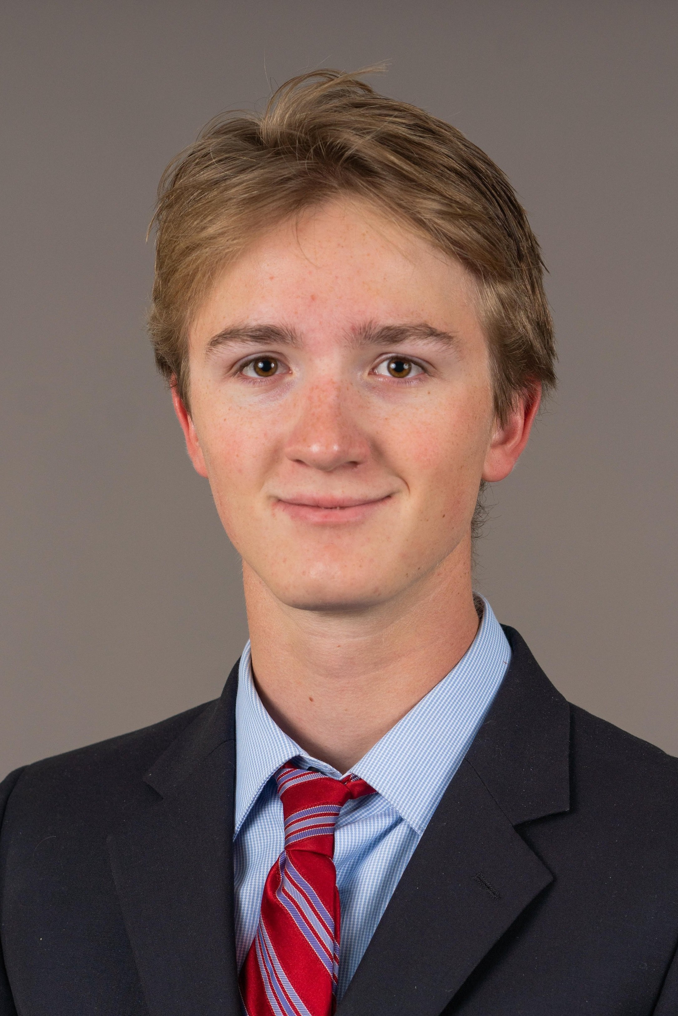 Young man wearing a dark suit, light blue dress shirt, red striped tie, and smiling against a plain background.